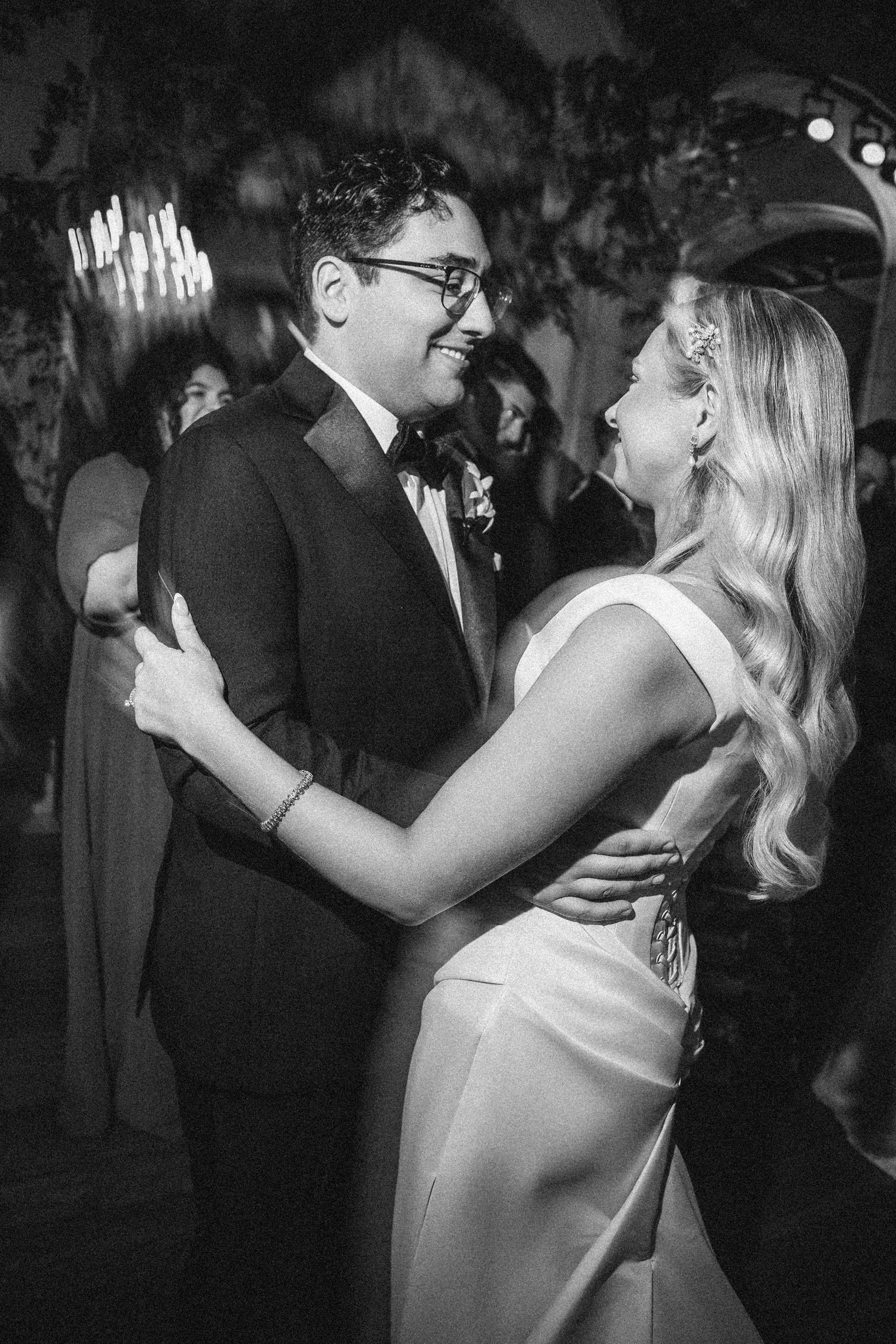 A couple dressed formally share a dance and smile at each other in a softly lit indoor setting, surrounded by other people, capturing the romantic ambiance of a Hempstead House at Sands Point Preserve wedding.