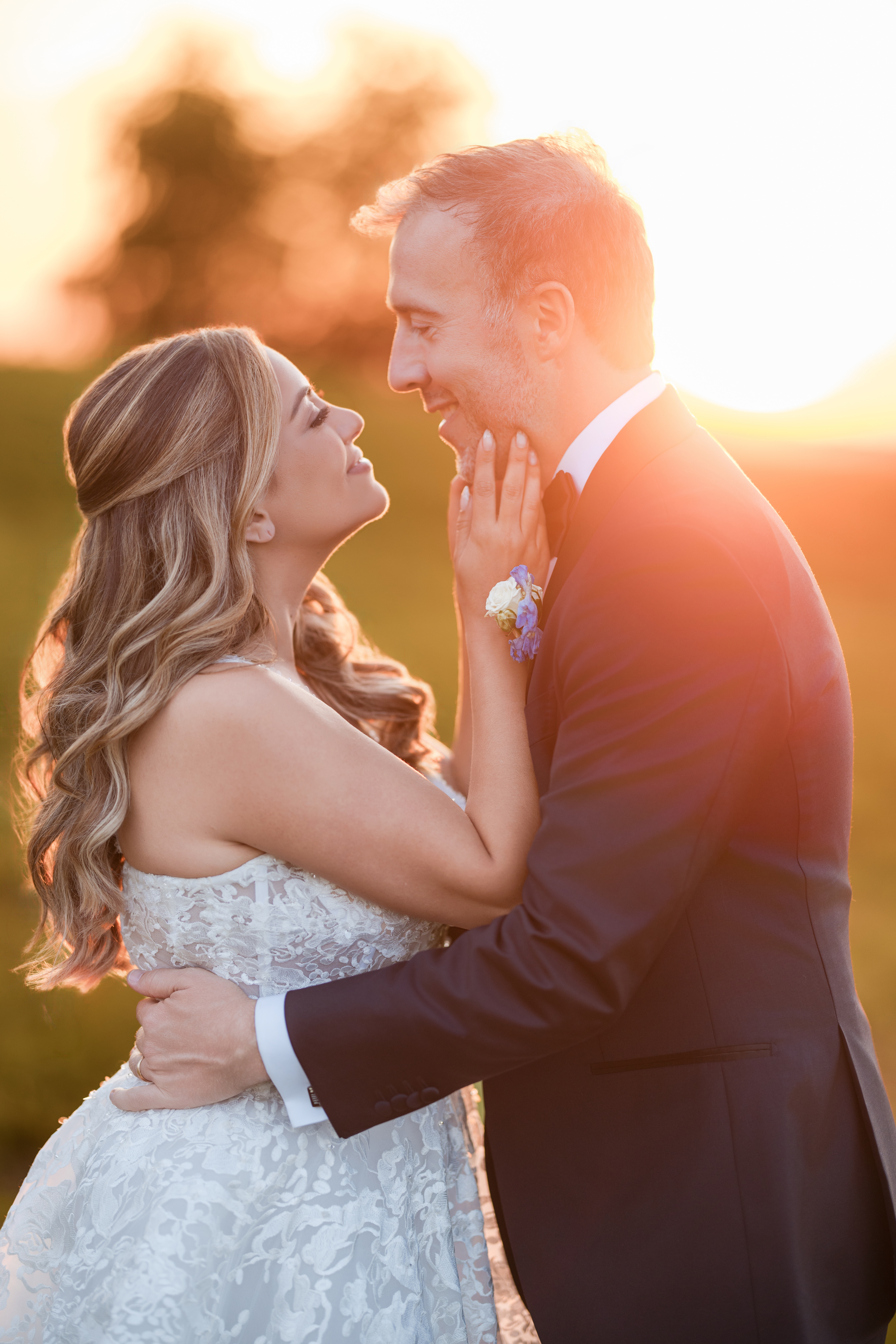 A bride and groom stand outdoors at sunset during their Regatta Place wedding, facing each other and smiling, with the bride’s hand gently touching the groom’s face.