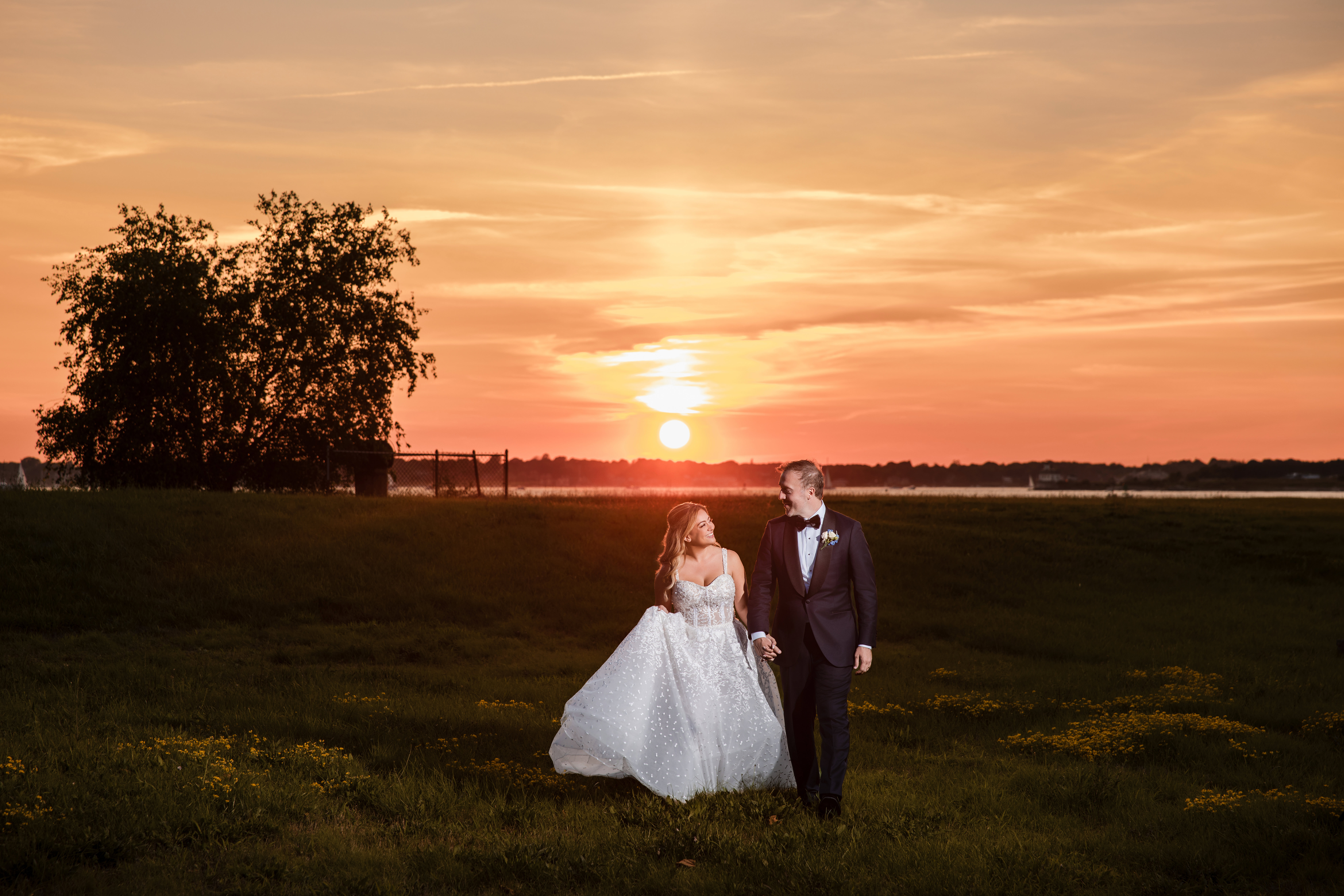 A bride and groom hold hands while walking in a grassy field at sunset, with the sun low on the horizon and trees in the background, capturing a romantic moment reminiscent of a Regatta Place wedding.