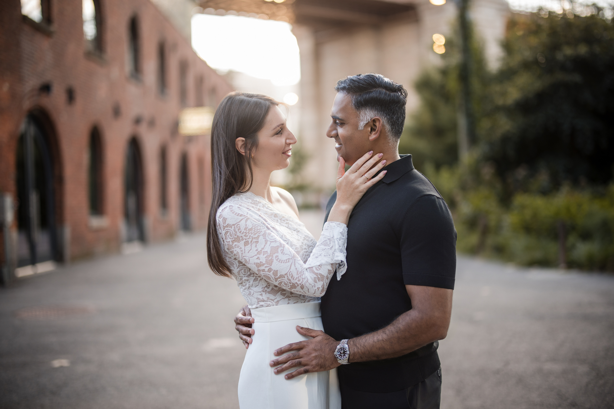 A woman in a white dress and a man in a black shirt stand outdoors during their DUMBO Engagement Session, facing each other closely and smiling, with one arm around each other. A woman in a white dress and a man in a black shirt stand outdoors during their DUMBO Engagement Session, facing each other closely and smiling, with one arm around each other.