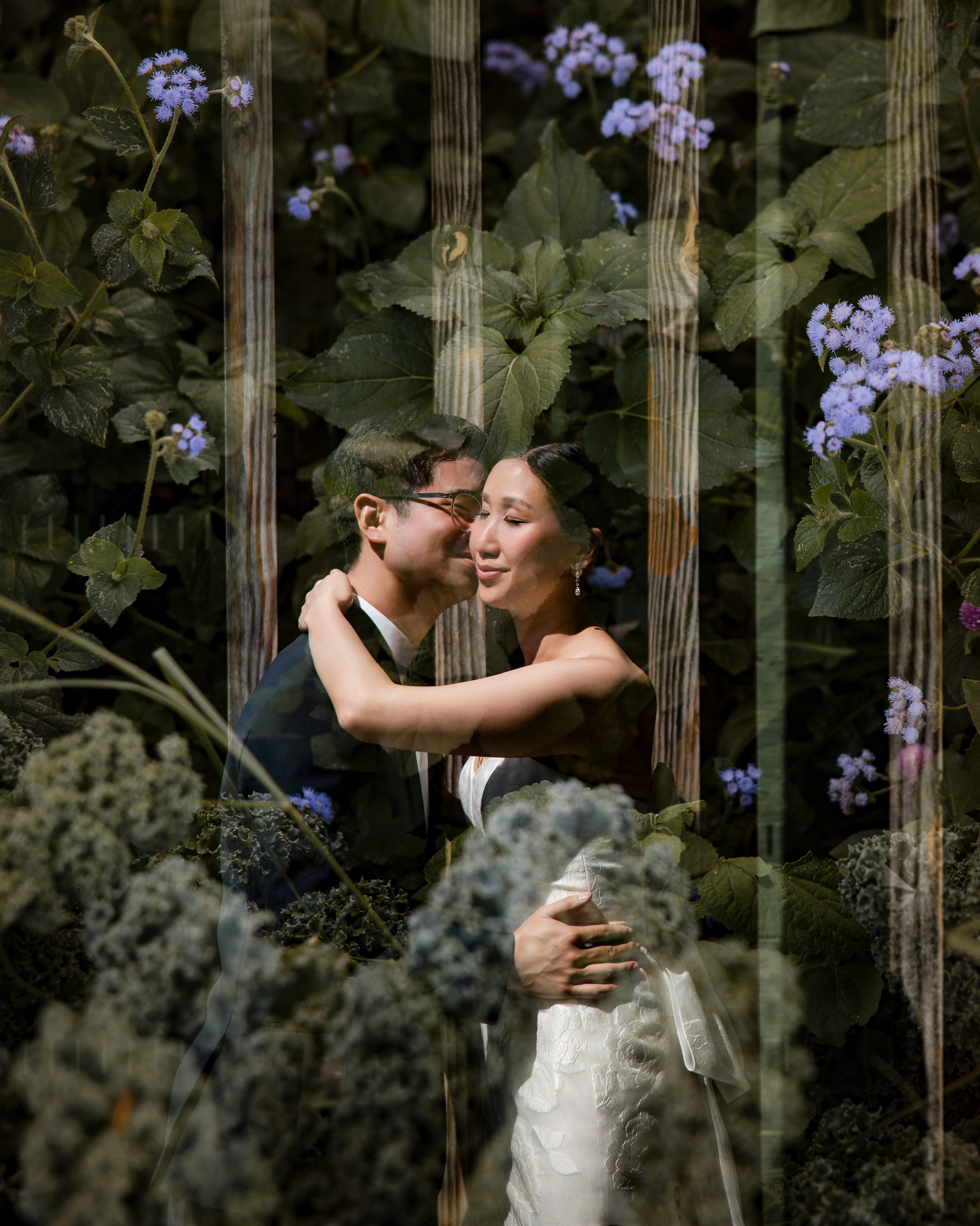 A couple in wedding attire embraces in front of leafy green foliage with purple flowers at their Brooklyn Botanic Garden Palm House wedding; vertical light streaks overlay the image.