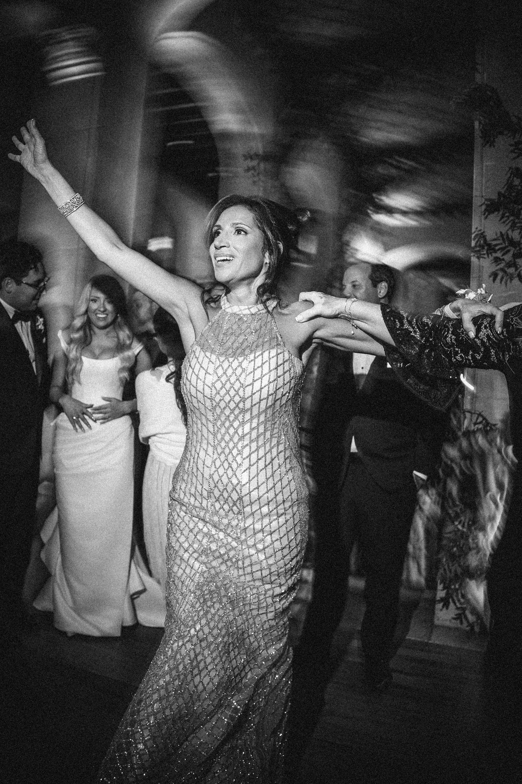 A woman in a patterned dress dances with arms raised at an indoor event, surrounded by people in formal attire at a Hempstead House at Sands Point Preserve wedding.