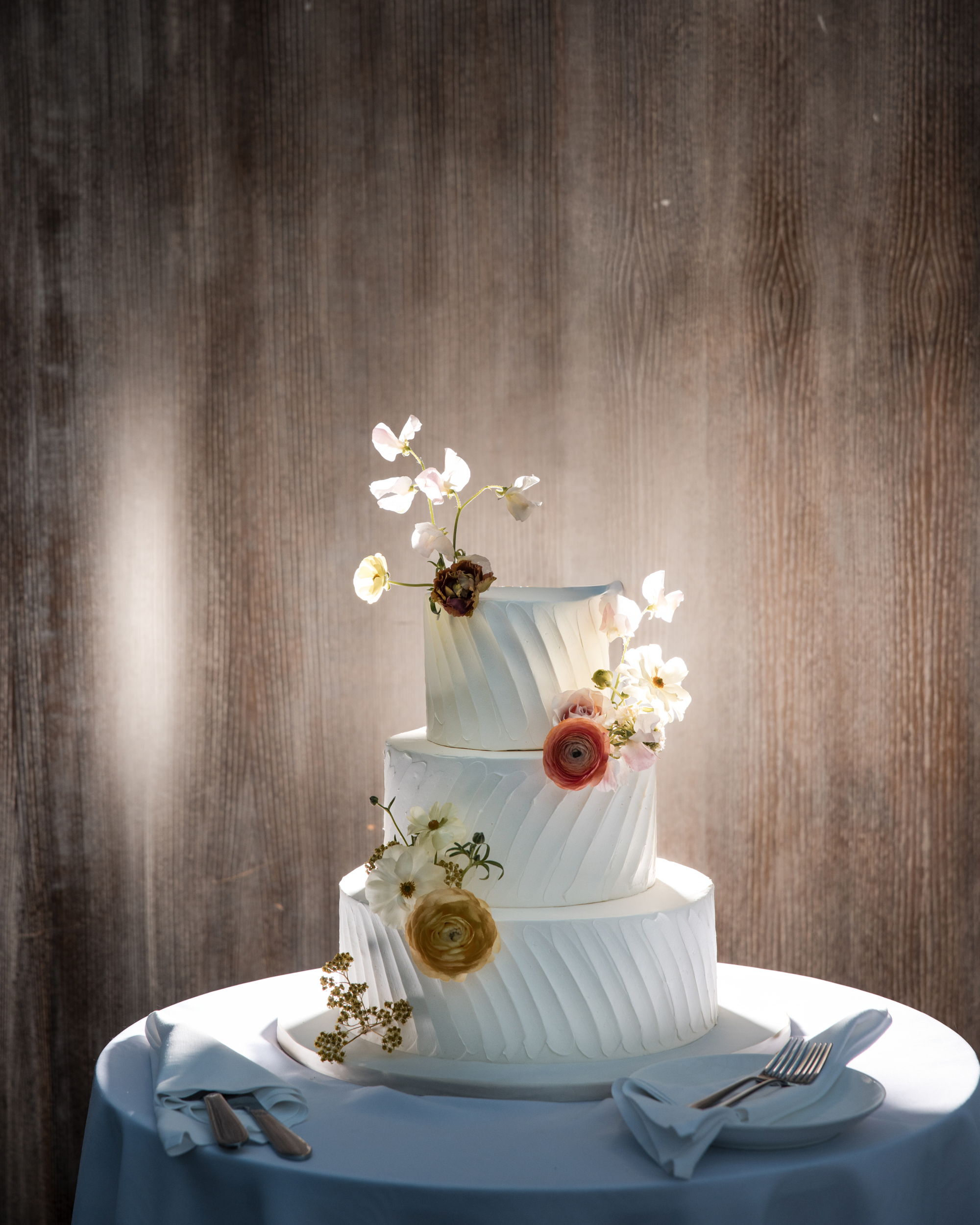 Three-tiered white cake with diagonal textured icing and pastel flowers, displayed on a round table with plates and utensils against a wooden background—perfect for an elegant Brooklyn Botanic Garden Palm House wedding.