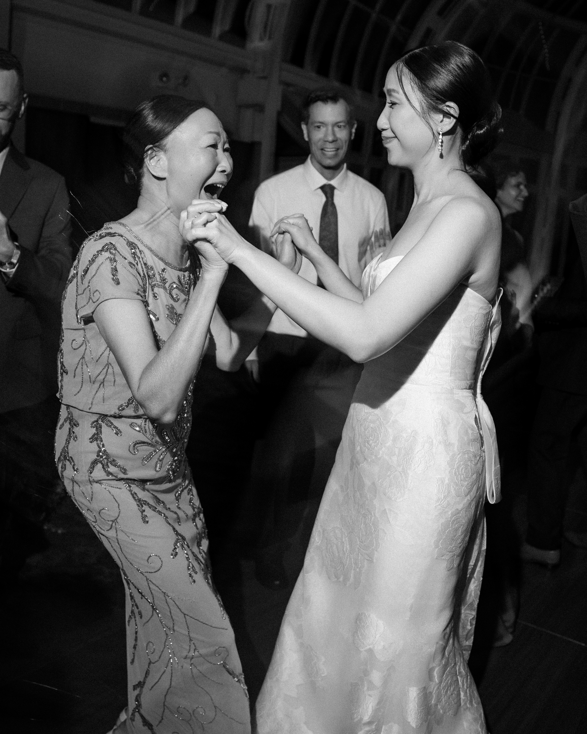 Two women, one a bride in a gown and one in a formal dress, dance together at an indoor event, their joy illuminated by the glass walls of the Brooklyn Botanic Garden Palm House wedding venue as others watch and smile in the background.