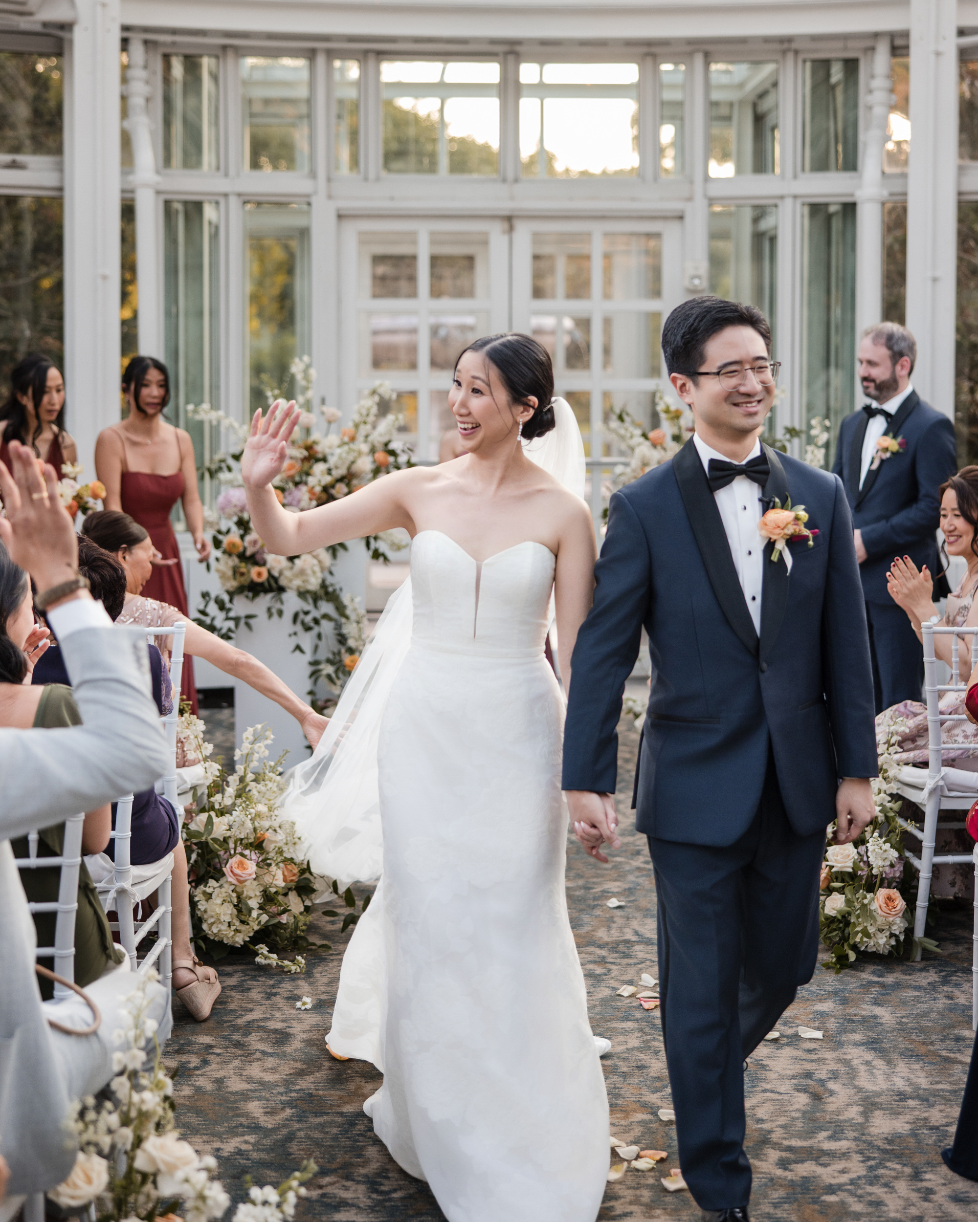 A bride and groom walk down the aisle, holding hands, smiling, and waving as guests seated on both sides applaud in the bright, flower-filled Brooklyn Botanic Garden Palm House wedding venue.
