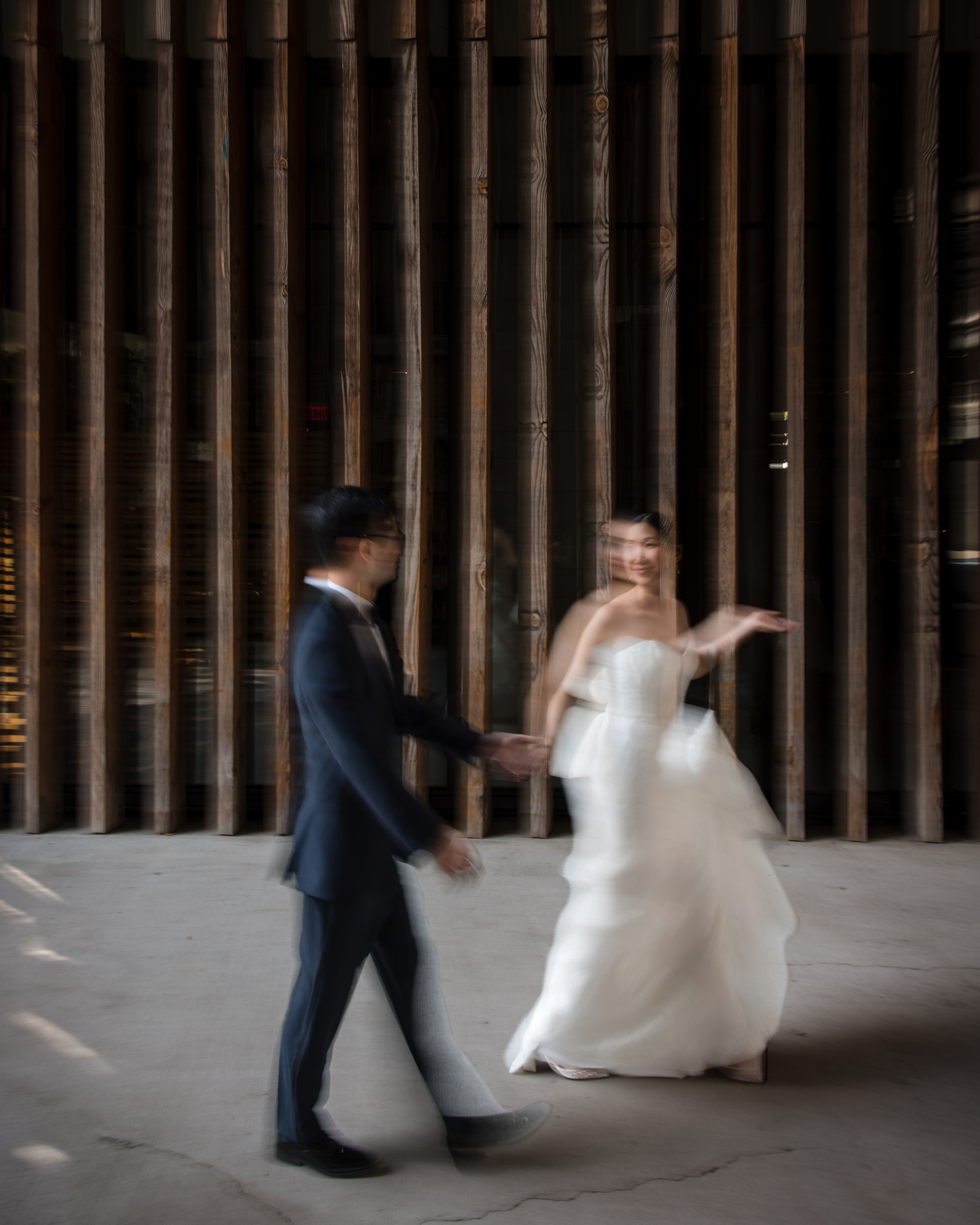 A bride in a white dress and a groom in a dark suit hold hands and move together in front of vertical wooden slats at their Brooklyn Botanic Garden Palm House wedding, creating a blurred motion effect.