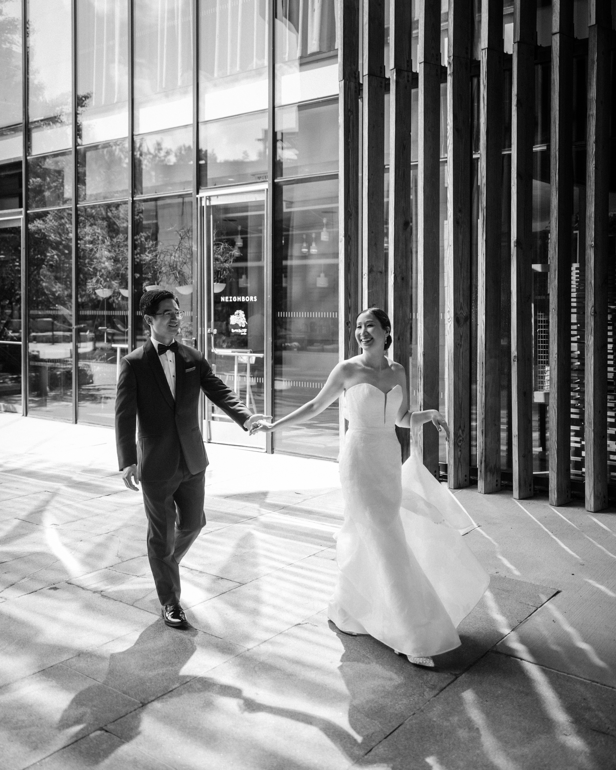 A bride and groom walk hand in hand outside the modern Brooklyn Botanic Garden Palm House. The groom wears a suit and bow tie; the bride wears a strapless gown. Sunlight creates patterns on the ground, adding to the charm of their Palm House wedding.
