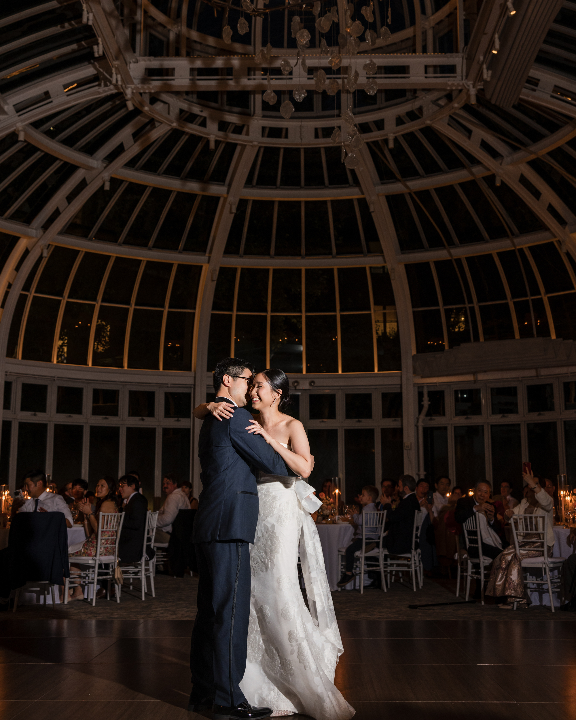 A bride and groom share their first dance in the elegant glass-domed Brooklyn Botanic Garden Palm House wedding venue, surrounded by seated guests and candlelit tables.