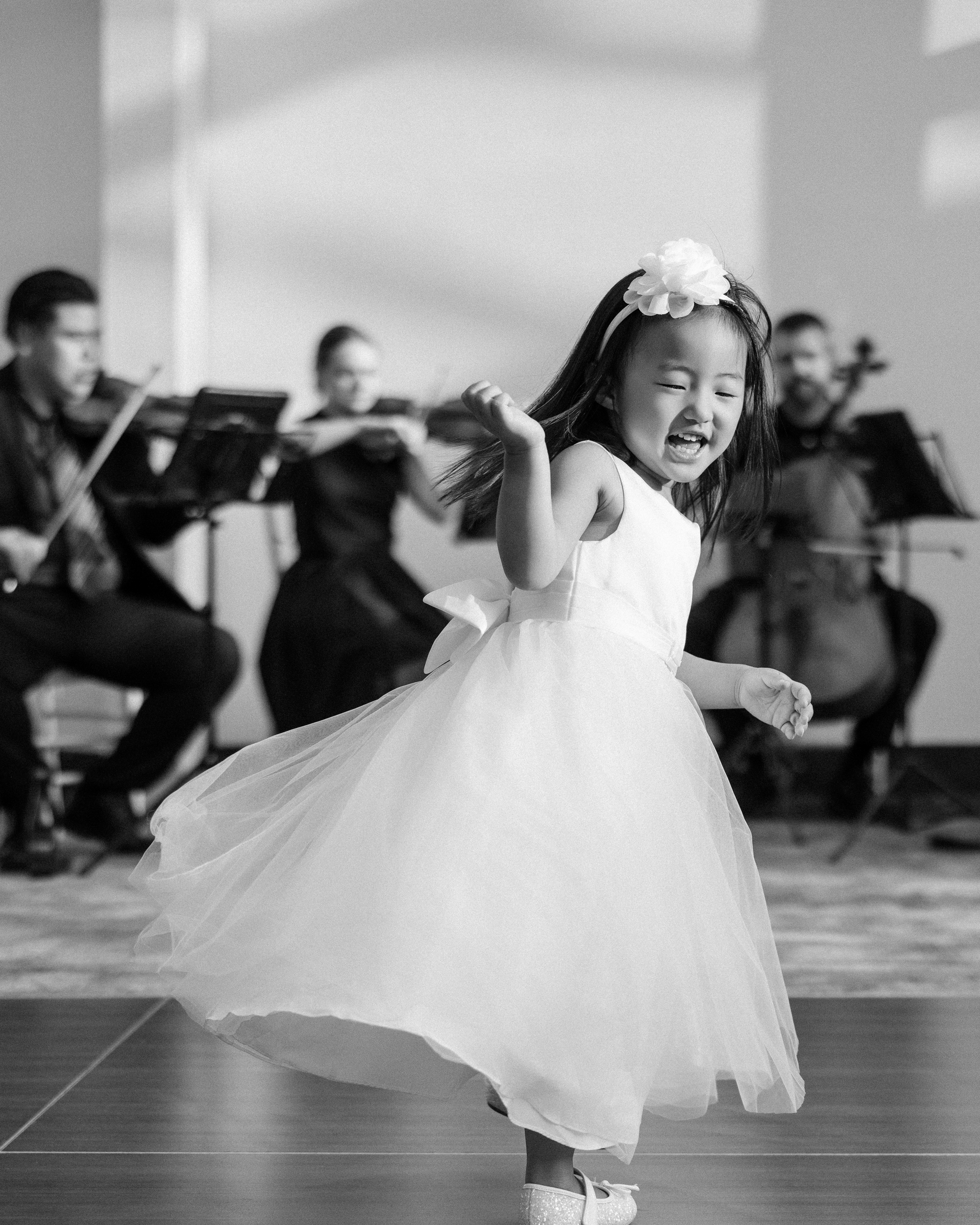 A young flower girl in a white dress dances joyfully in front of a seated string quartet at a Brooklyn Botanic Garden Palm House wedding, with sunlight casting shadows on the wall behind.
