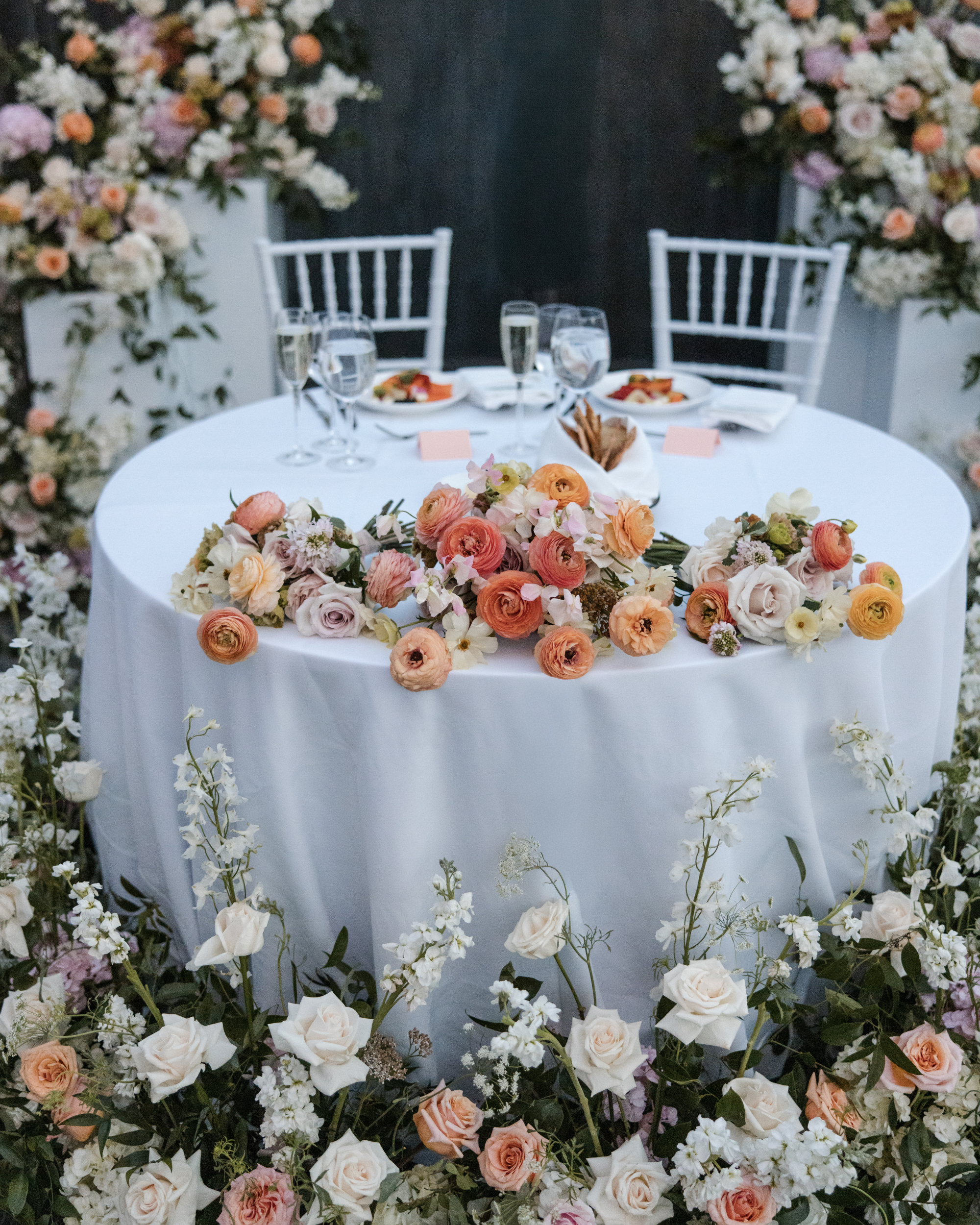 A round table with a white tablecloth is decorated with pastel flower arrangements and surrounded by white chairs and additional floral displays, evoking the elegance of a Brooklyn Botanic Garden Palm House wedding. Plates and glasses are set on the table.