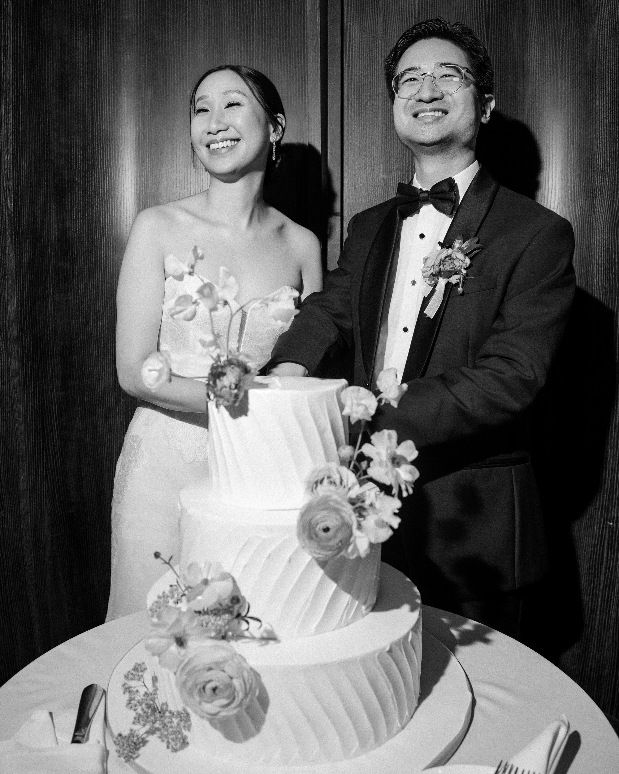 A bride and groom, both smiling, stand together as they cut a tiered wedding cake decorated with flowers during their elegant Brooklyn Botanic Garden Palm House wedding.
