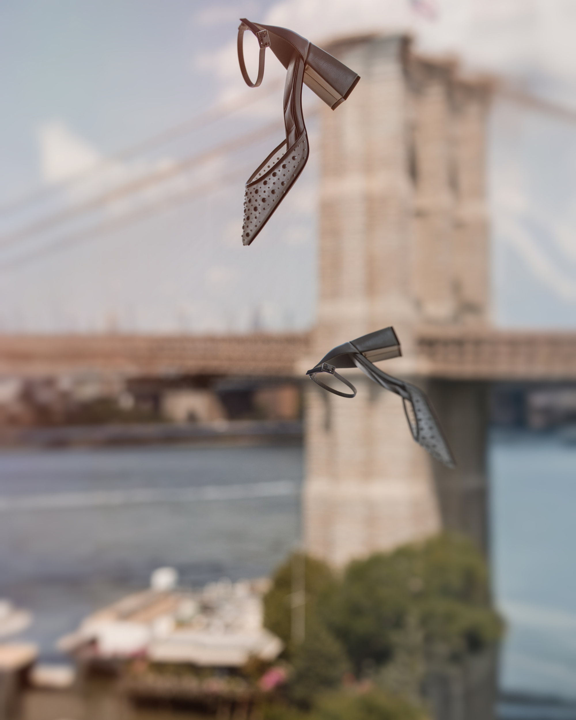 Two heeled sandals are suspended in mid-air with the Brooklyn Bridge and East River visible in the blurred background on a clear day, evoking the elegance of a Brooklyn Botanic Garden Palm House wedding.