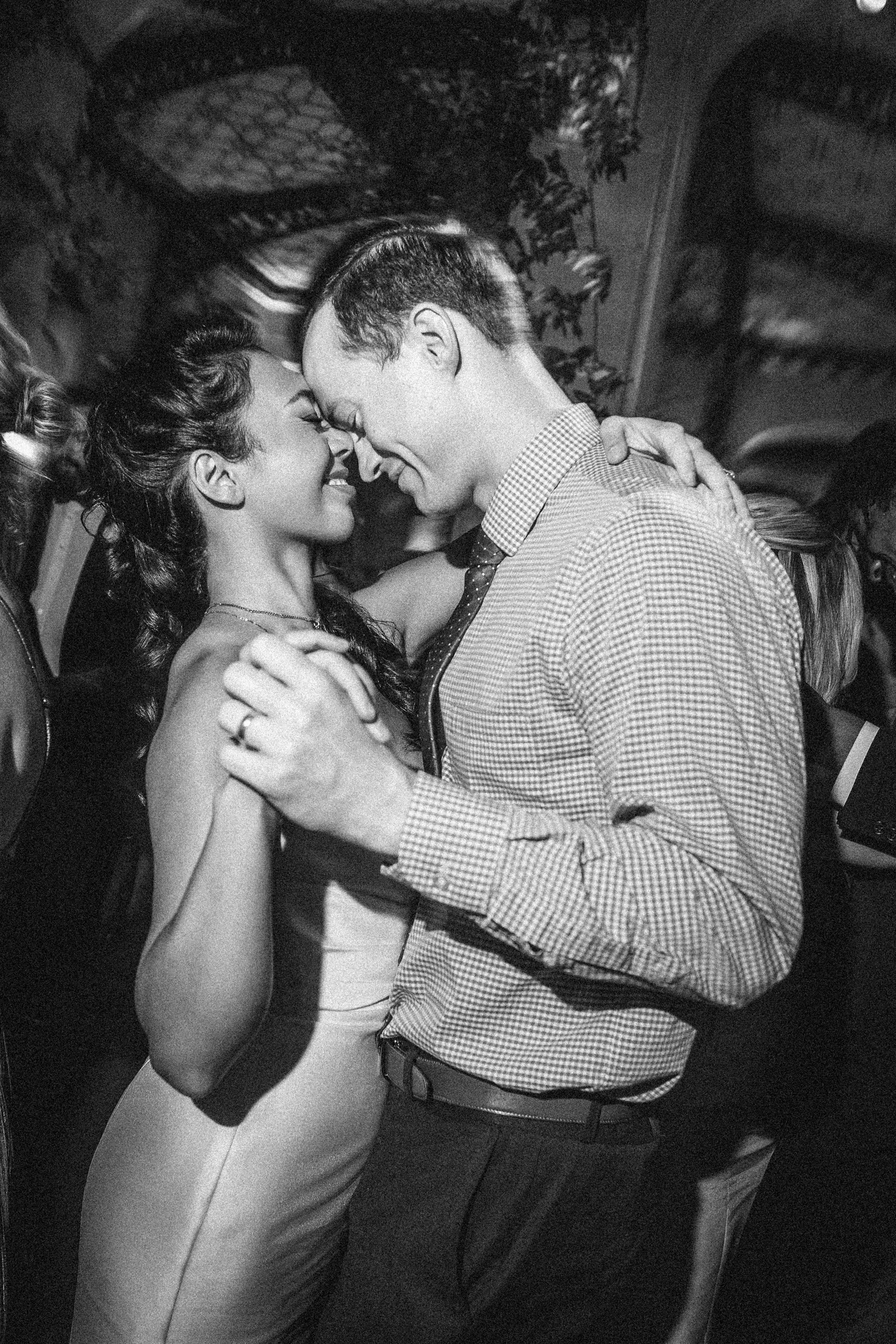 A couple dances closely together, smiling and holding hands, in a dimly lit room with other people in the background at a beautiful Hempstead House at Sands Point Preserve wedding.