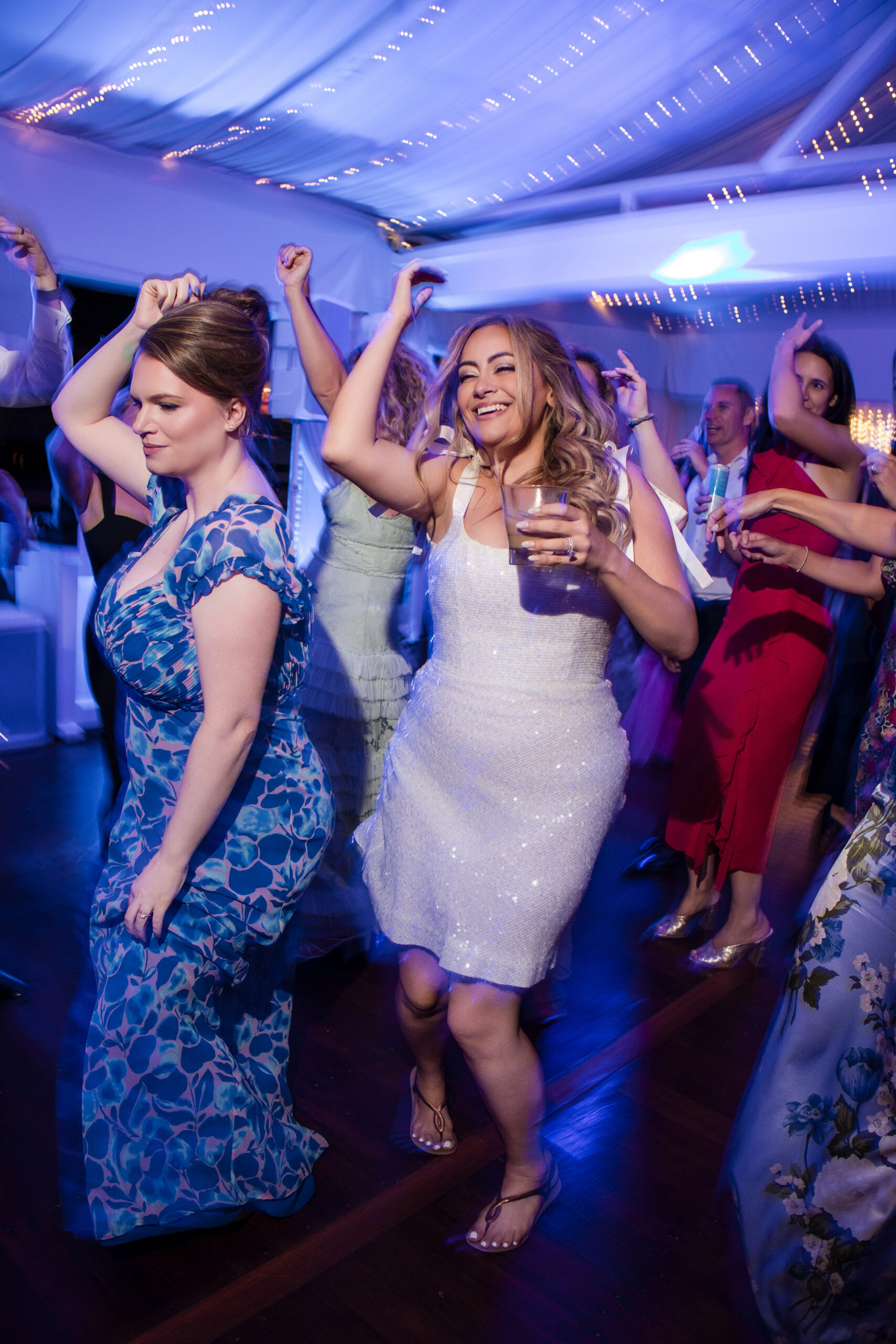 People dance energetically at an indoor Regatta Place wedding under string lights. A woman in a white dress holds a drink and smiles while dancing among others in colorful outfits.