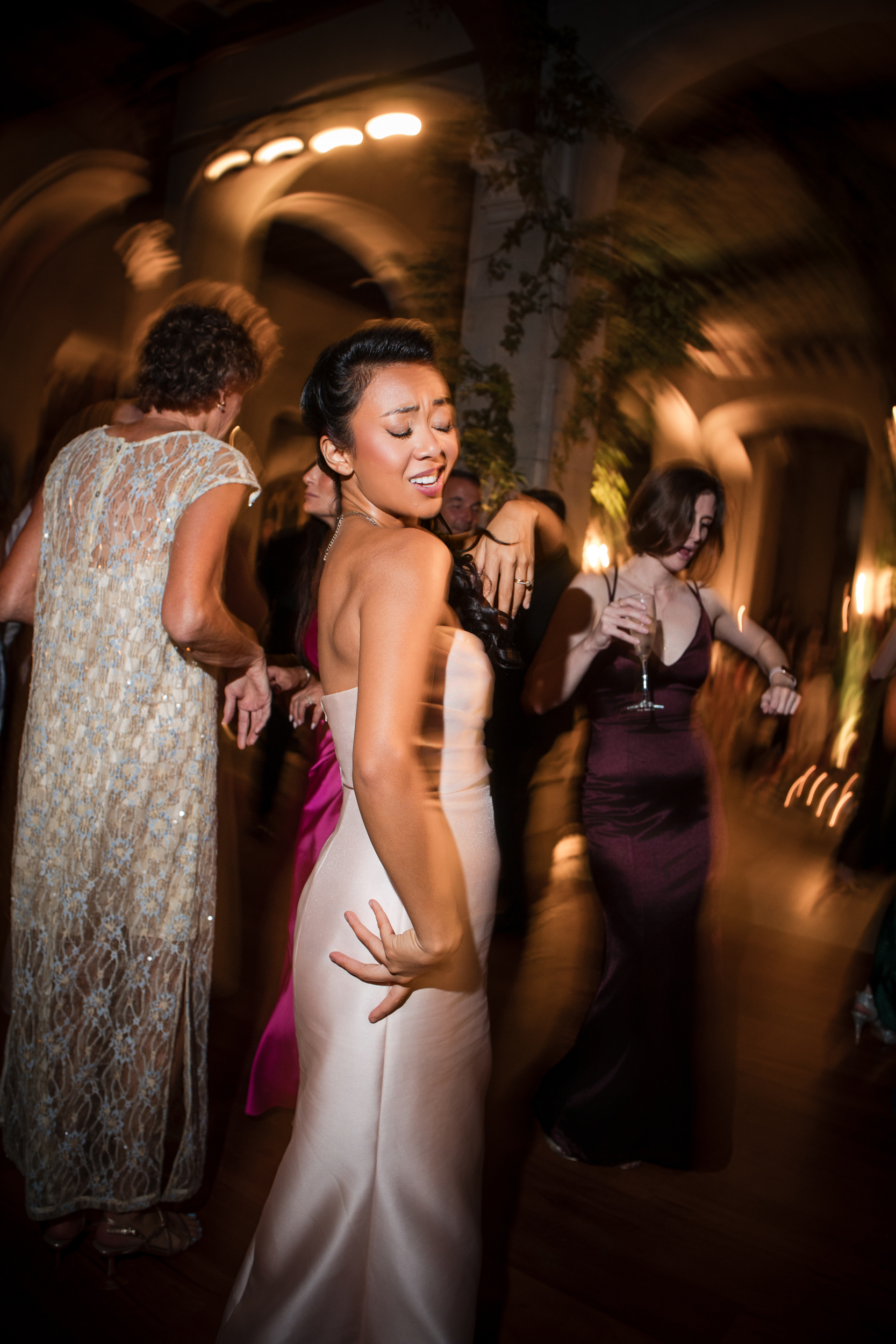 A woman in a light dress dances energetically at an indoor event, surrounded by other guests in formal attire under warm lighting at a beautiful Hempstead House at Sands Point Preserve wedding.