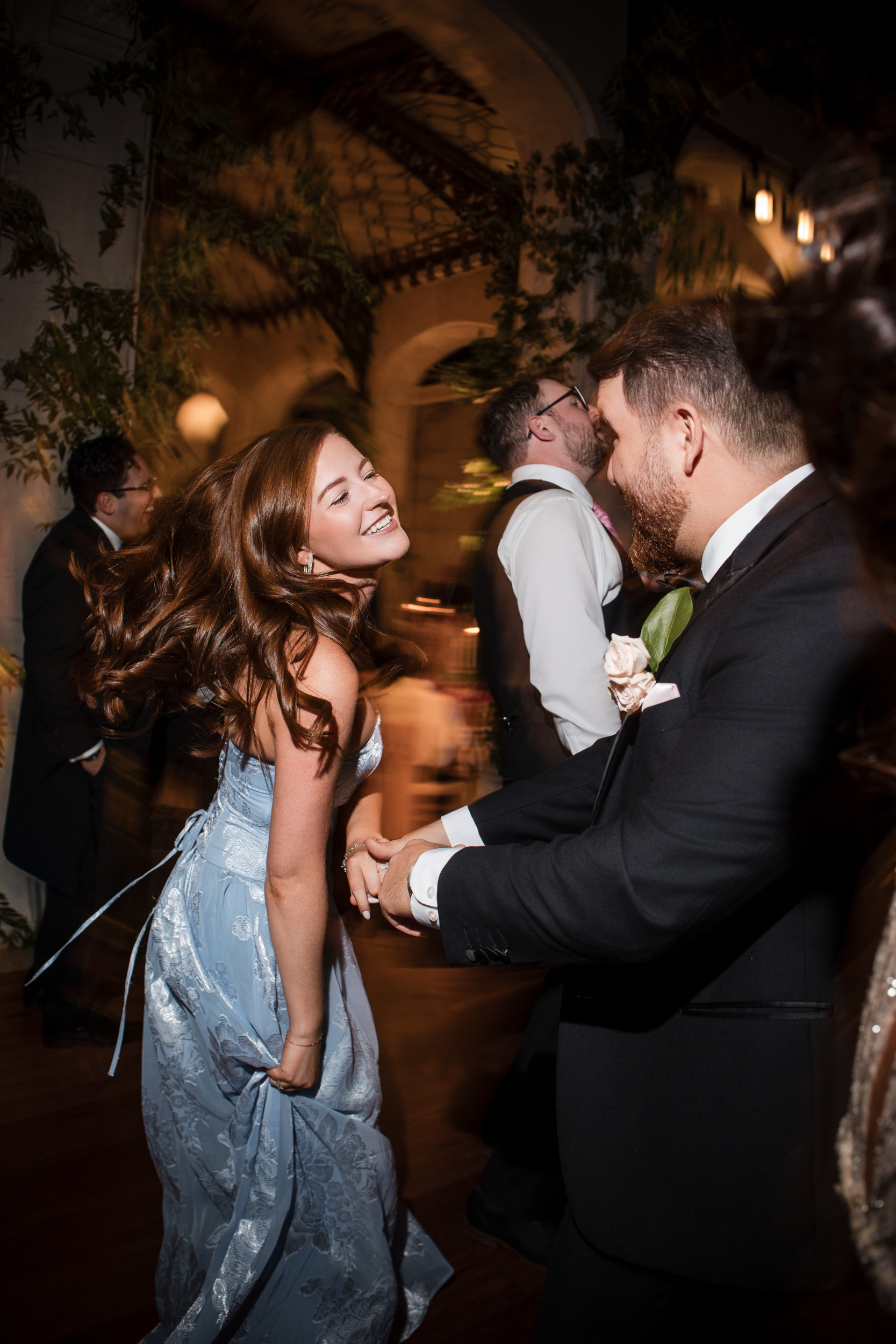 A woman in a light blue dress dances joyfully with a man in a suit at an indoor event, surrounded by other people, capturing the elegance of a Hempstead House at Sands Point Preserve wedding.