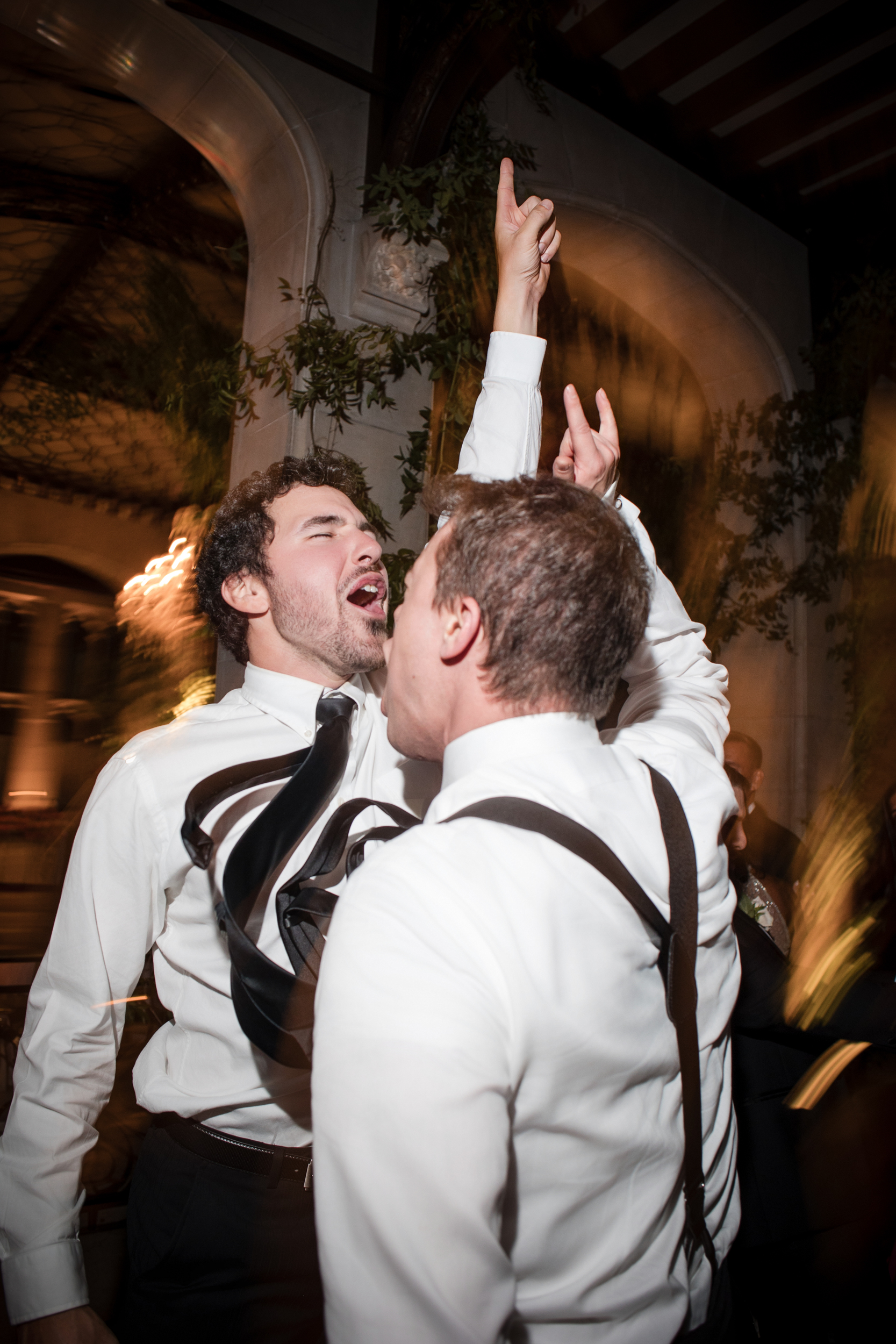 Two men in dress shirts and suspenders enthusiastically sing and gesture with raised arms at an indoor event, capturing the lively spirit of a Hempstead House at Sands Point Preserve wedding.