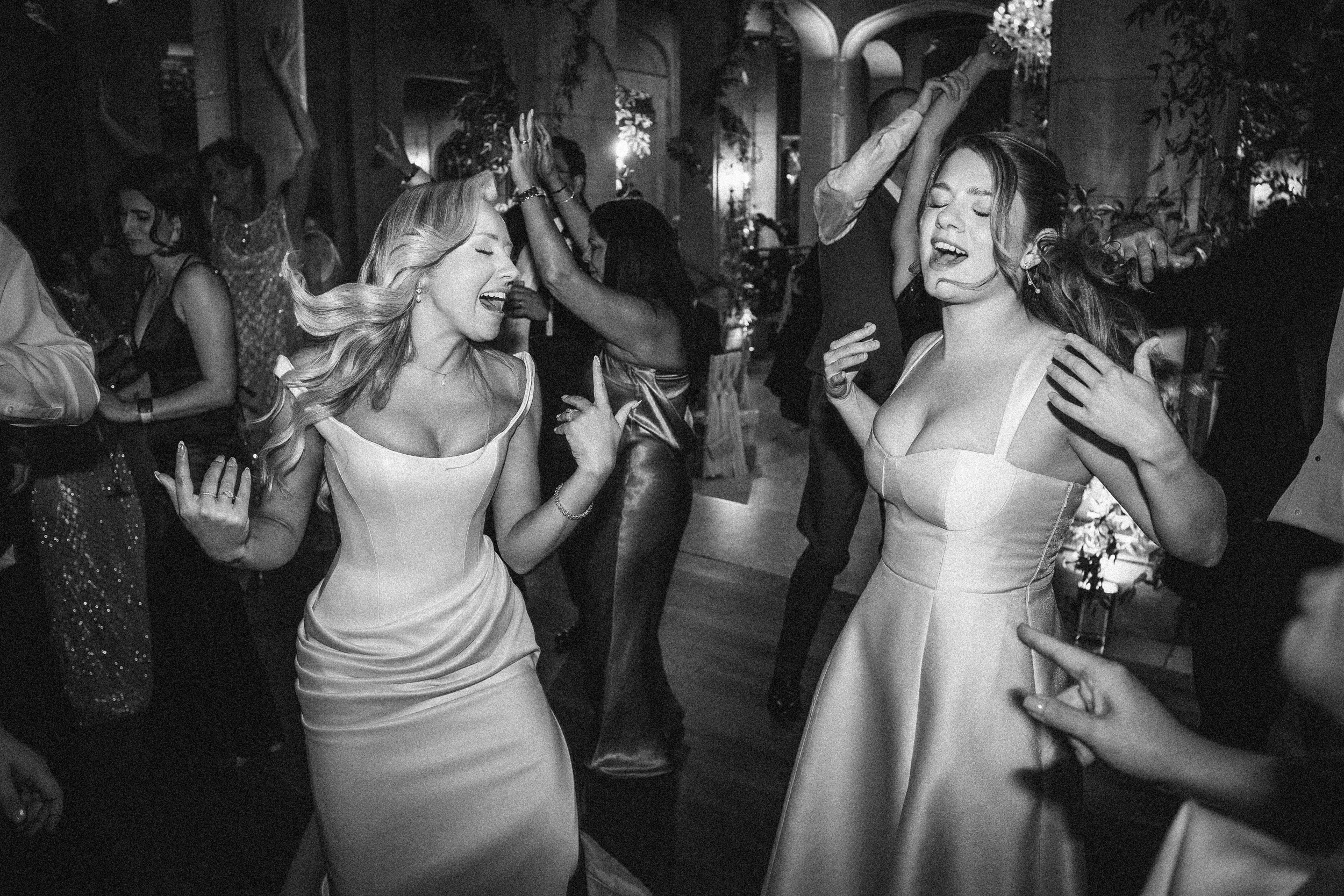 Two women in formal dresses dance with eyes closed and hands raised at an indoor event, surrounded by other people, capturing the elegant spirit of a Hempstead House at Sands Point Preserve wedding. Two women in formal dresses dance with eyes closed and hands raised at an indoor event, surrounded by other people, capturing the elegant spirit of a Hempstead House at Sands Point Preserve wedding.