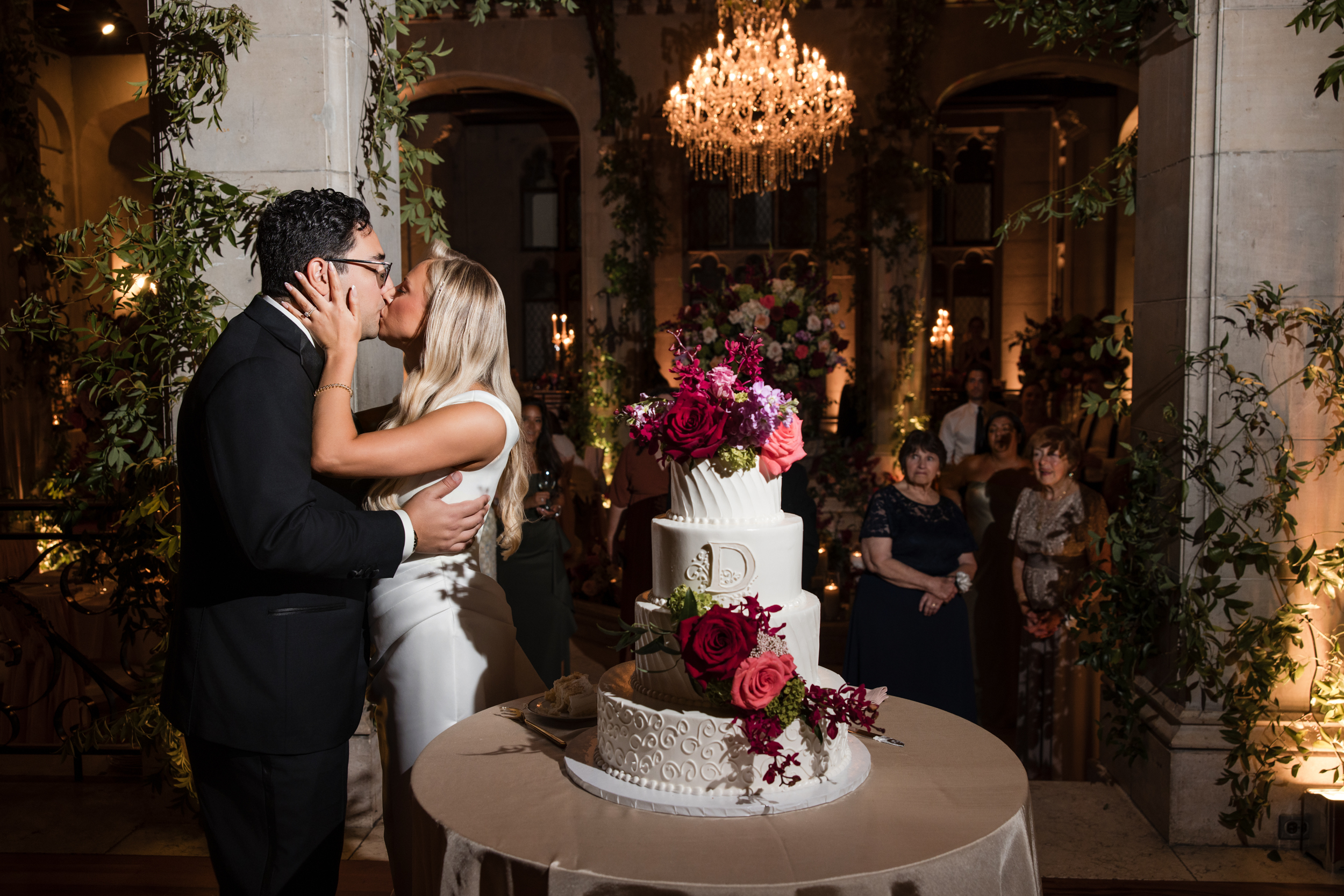 A bride and groom kiss in front of a wedding cake at an indoor reception at Hempstead House at Sands Point Preserve, with guests observing in the background under a chandelier. A bride and groom kiss in front of a wedding cake at an indoor reception at Hempstead House at Sands Point Preserve, with guests observing in the background under a chandelier.