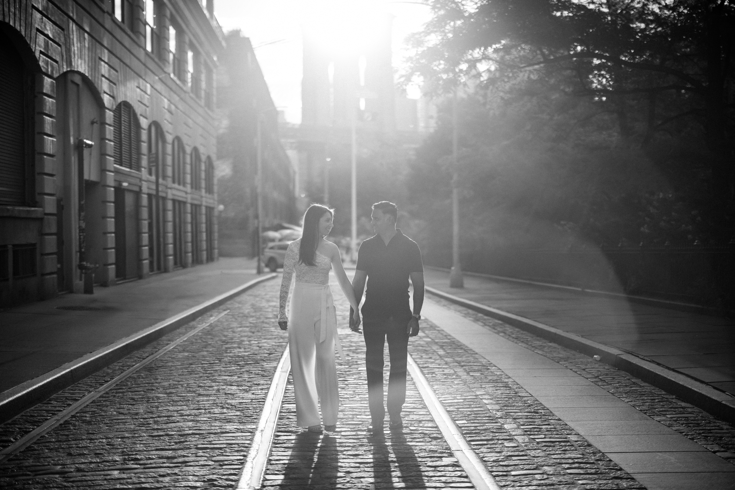 A couple walks hand in hand on a cobblestone street with sunlight streaming behind them, surrounded by buildings and trees—capturing the magic of a DUMBO Engagement Session. A couple walks hand in hand on a cobblestone street with sunlight streaming behind them, surrounded by buildings and trees—capturing the magic of a DUMBO Engagement Session.