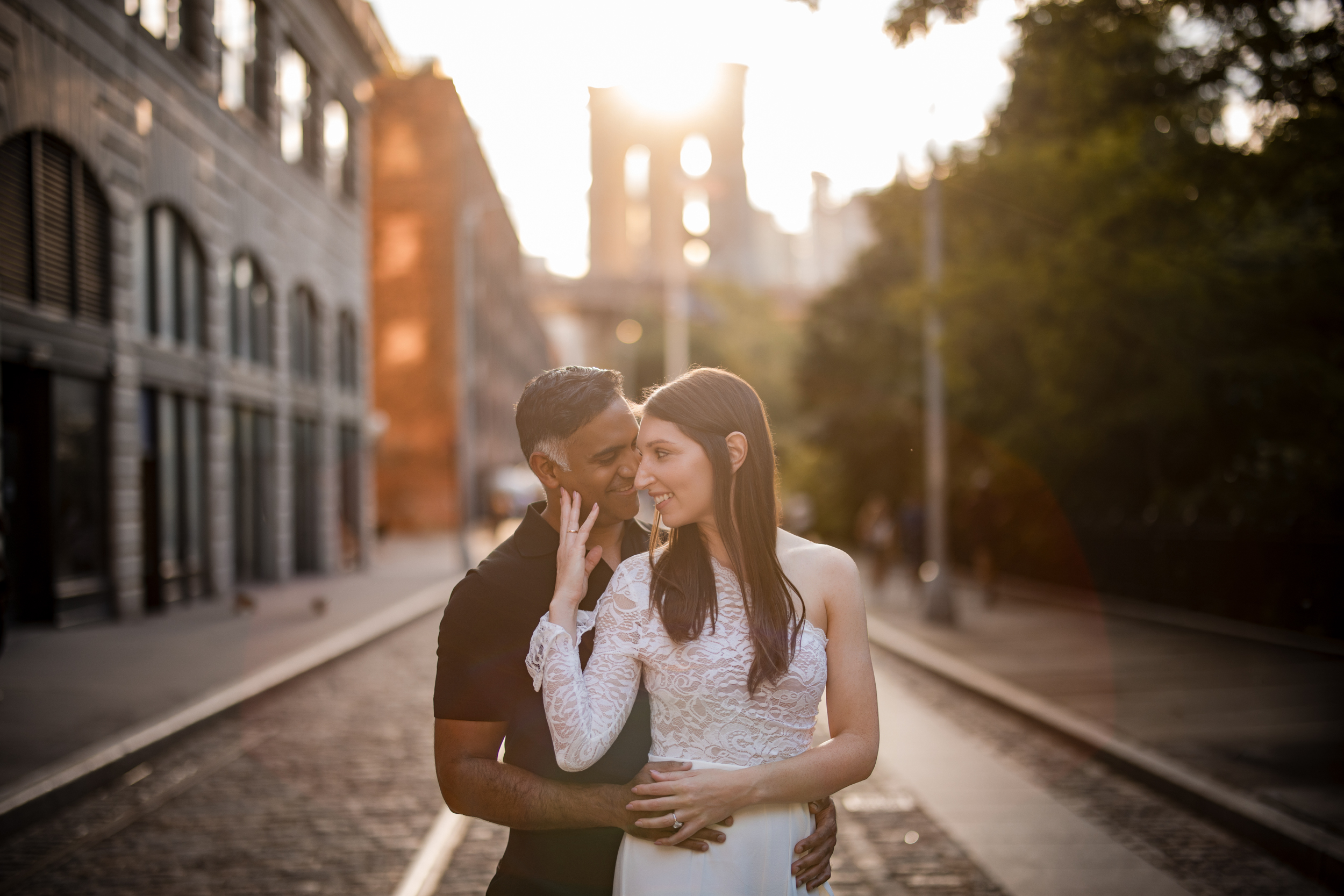 A couple stands close together on a cobblestone street at sunset with the Brooklyn Bridge visible in the background, capturing the magic of a DUMBO Engagement Session. A couple stands close together on a cobblestone street at sunset with the Brooklyn Bridge visible in the background, capturing the magic of a DUMBO Engagement Session.