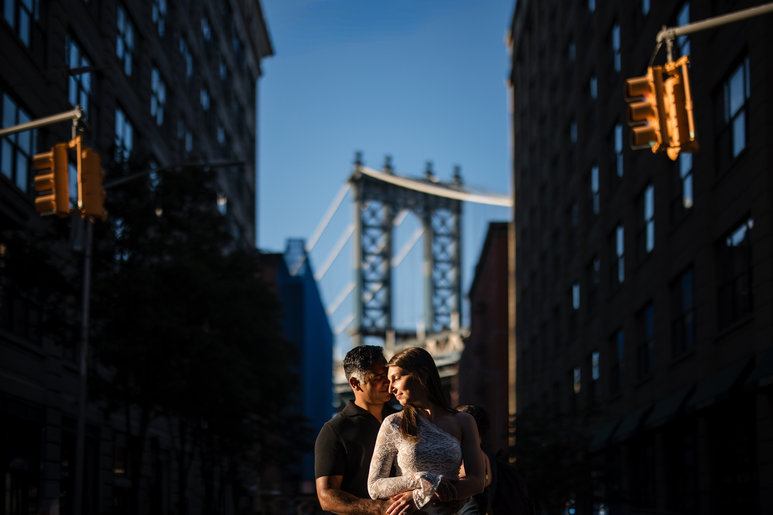 A couple stands closely together in the middle of a city street during their DUMBO Engagement Session, with a bridge and tall buildings in the background under bright sunlight. A couple stands closely together in the middle of a city street during their DUMBO Engagement Session, with a bridge and tall buildings in the background under bright sunlight.