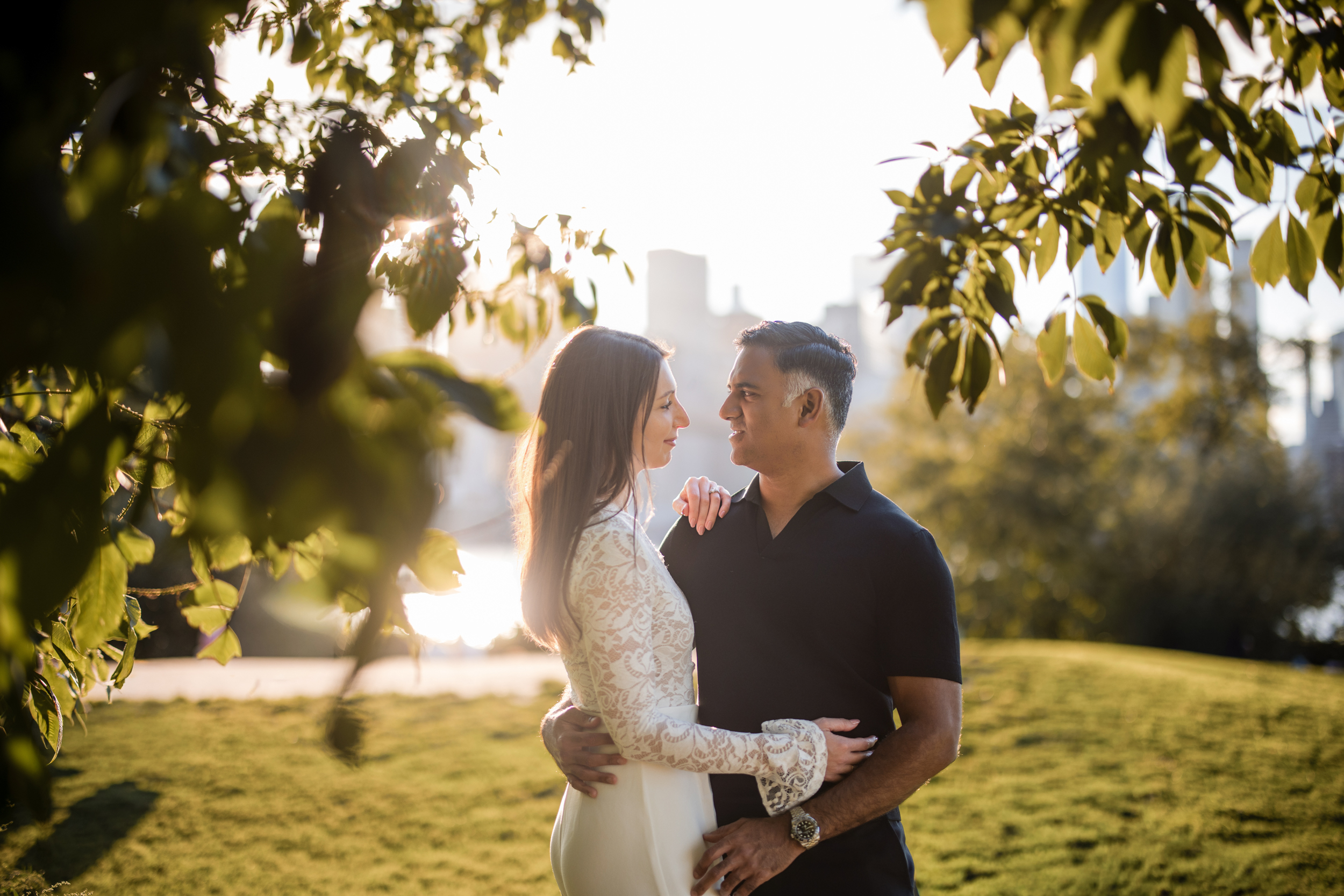 A woman in a white dress and a man in a black shirt stand close together outdoors, looking at each other and smiling, with greenery and sunlight in the background, capturing the joyful intimacy of their DUMBO engagement session. A woman in a white dress and a man in a black shirt stand close together outdoors, looking at each other and smiling, with greenery and sunlight in the background, capturing the joyful intimacy of their DUMBO engagement session.