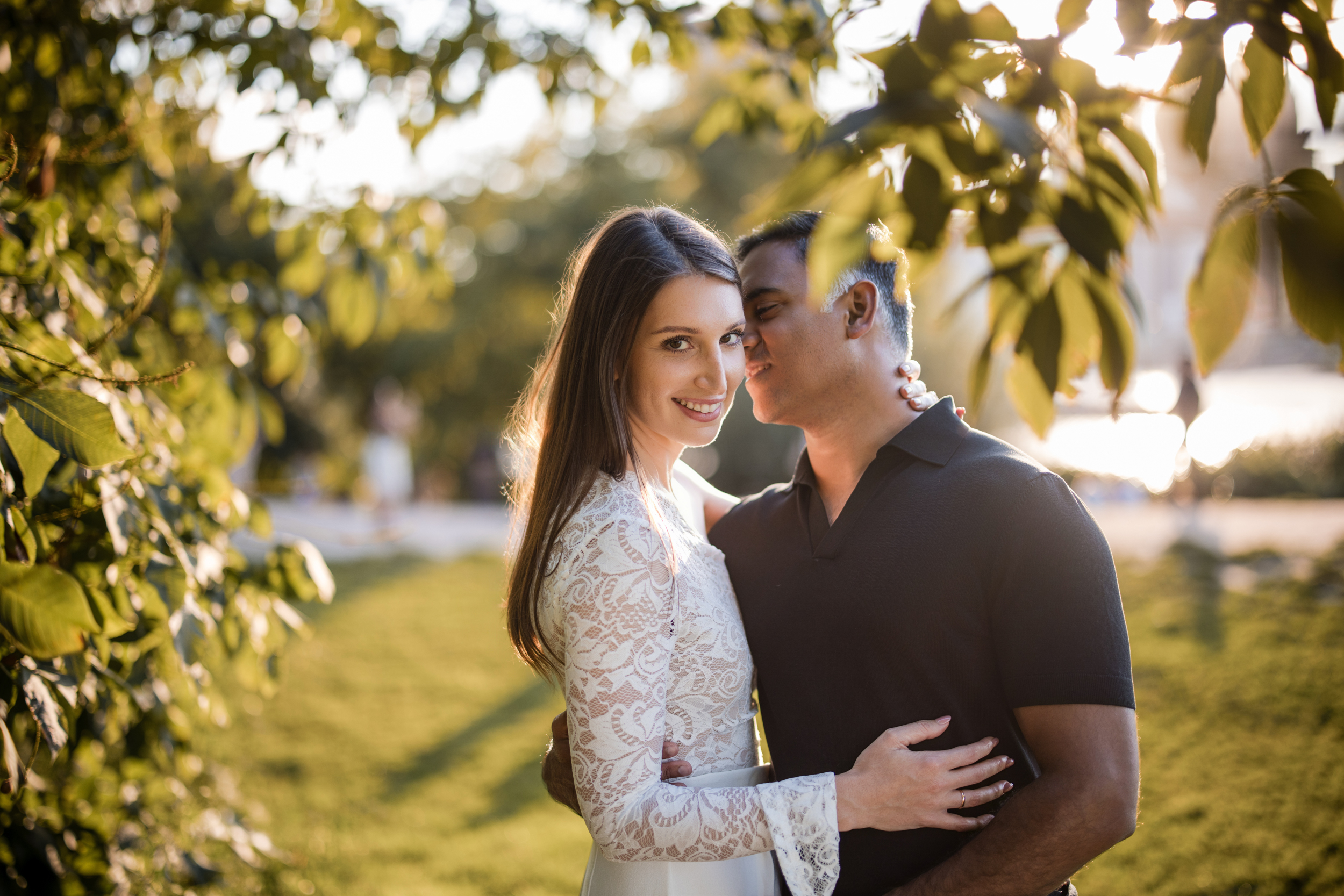 A woman in a white lace dress smiles at the camera while a man in a black shirt hugs and kisses her cheek during their sunlit DUMBO Engagement Session, surrounded by lush greenery.