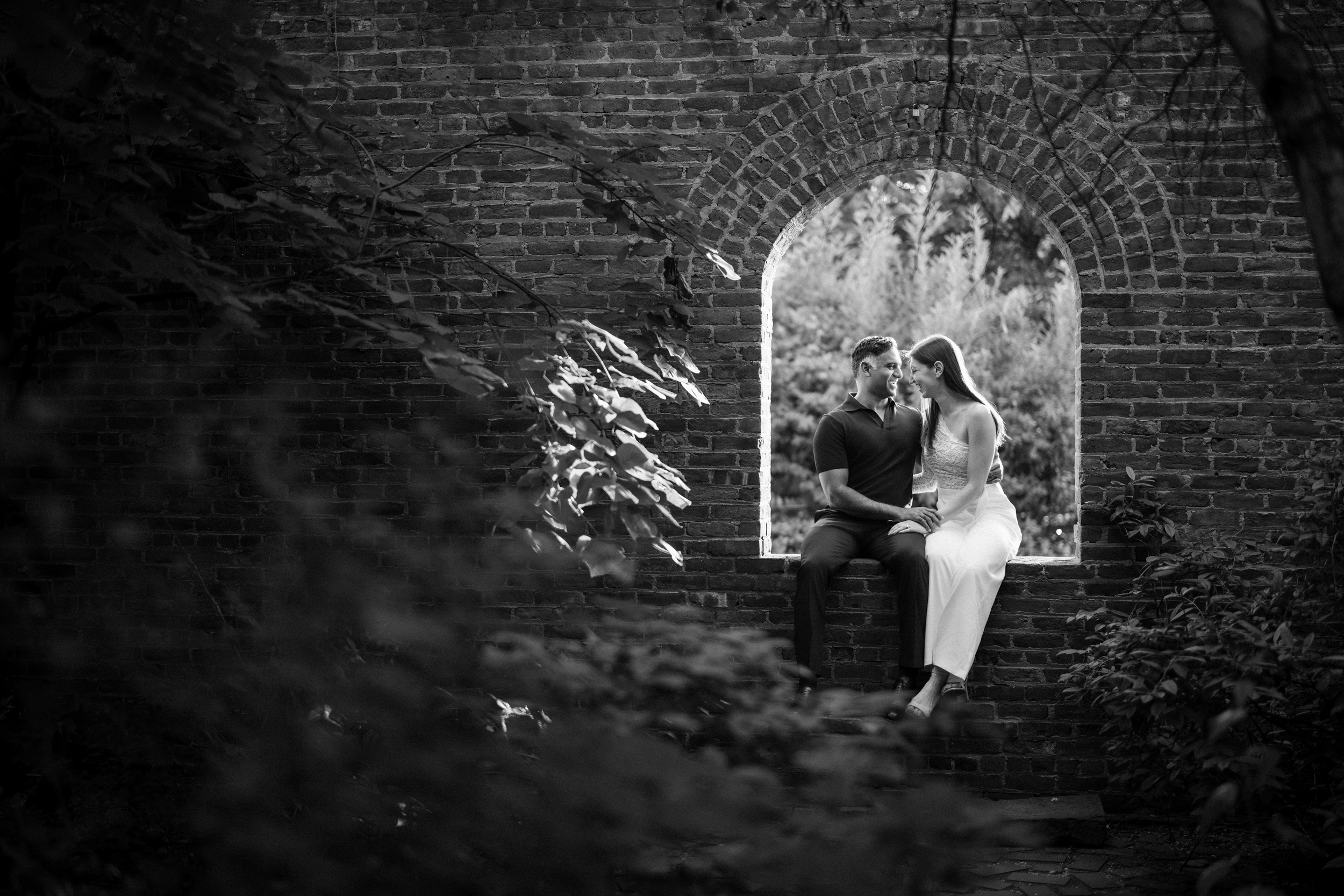 A couple sits together in a stone archway, surrounded by foliage during their DUMBO engagement session. The photo is black and white, with soft natural light illuminating the pair. A couple sits together in a stone archway, surrounded by foliage during their DUMBO engagement session. The photo is black and white, with soft natural light illuminating the pair.