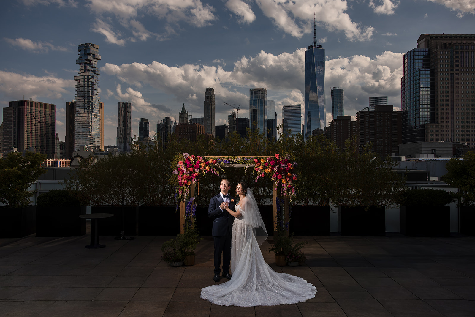 A bride and groom stand under a floral arch on an NYC rooftop wedding venue, with a city skyline of tall buildings and clouds in the background.