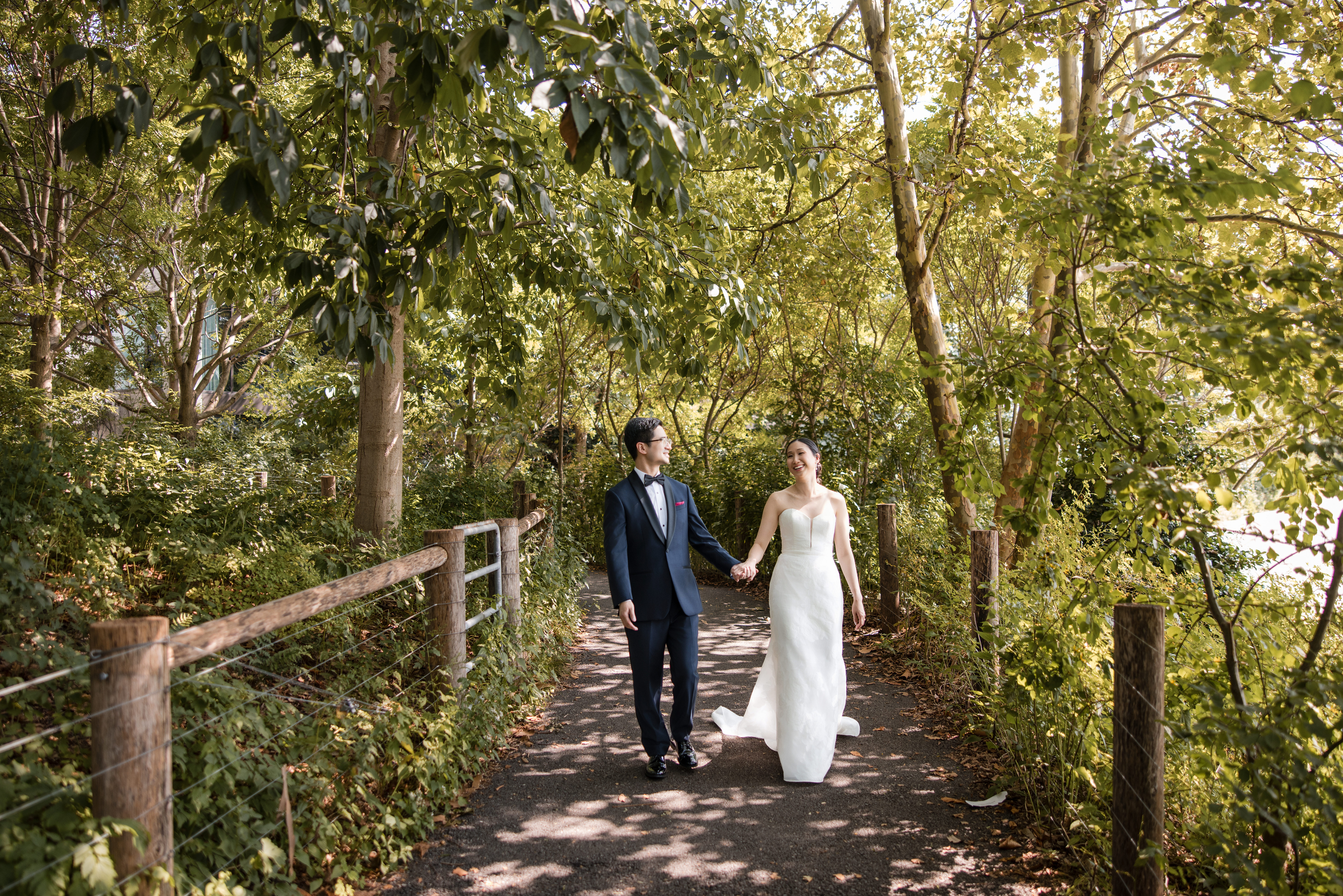 A couple in formal wedding attire walks hand in hand down a shaded path surrounded by green trees and foliage on a sunny day, capturing the romantic essence of a Brooklyn Botanic Garden Palm House wedding.