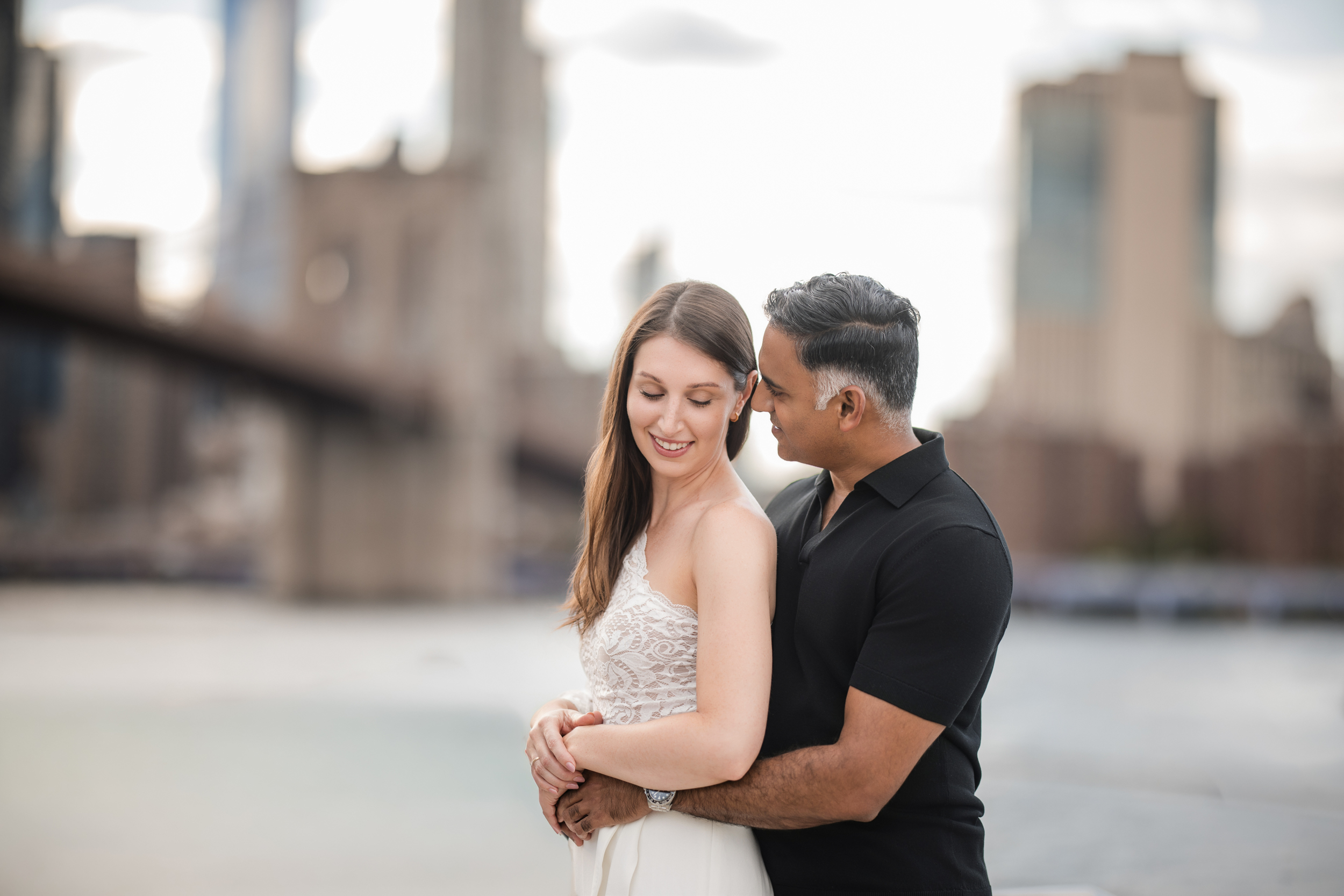 A man in a black shirt embraces a smiling woman in a white dress outdoors during their DUMBO Engagement Session, with blurred city buildings and a bridge in the background. A man in a black shirt embraces a smiling woman in a white dress outdoors during their DUMBO Engagement Session, with blurred city buildings and a bridge in the background.