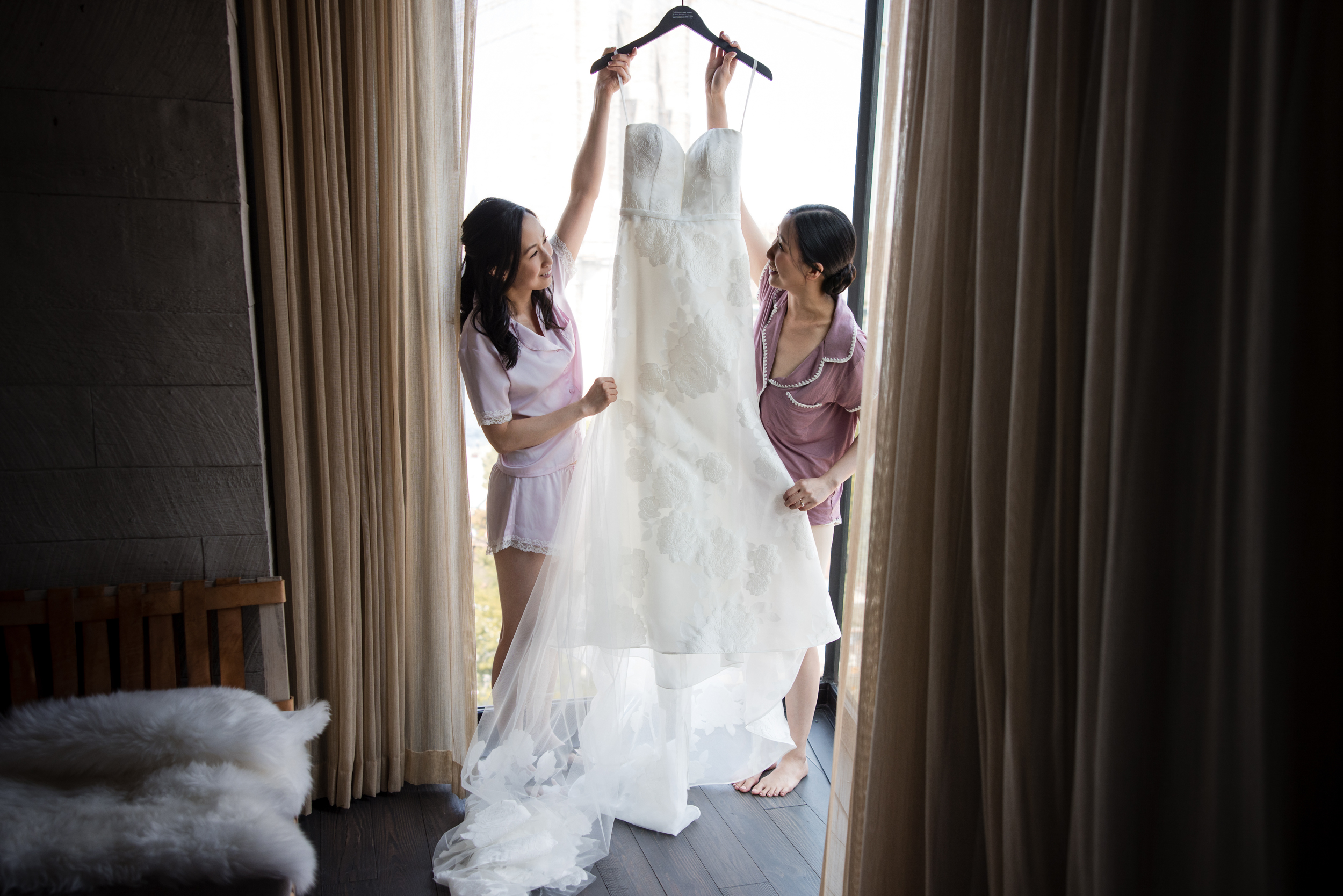 Two women in pajamas hold up a white wedding dress on a hanger by a large window with light curtains, preparing for a stunning Brooklyn Botanic Garden Palm House wedding. Two women in pajamas hold up a white wedding dress on a hanger by a large window with light curtains, preparing for a stunning Brooklyn Botanic Garden Palm House wedding.