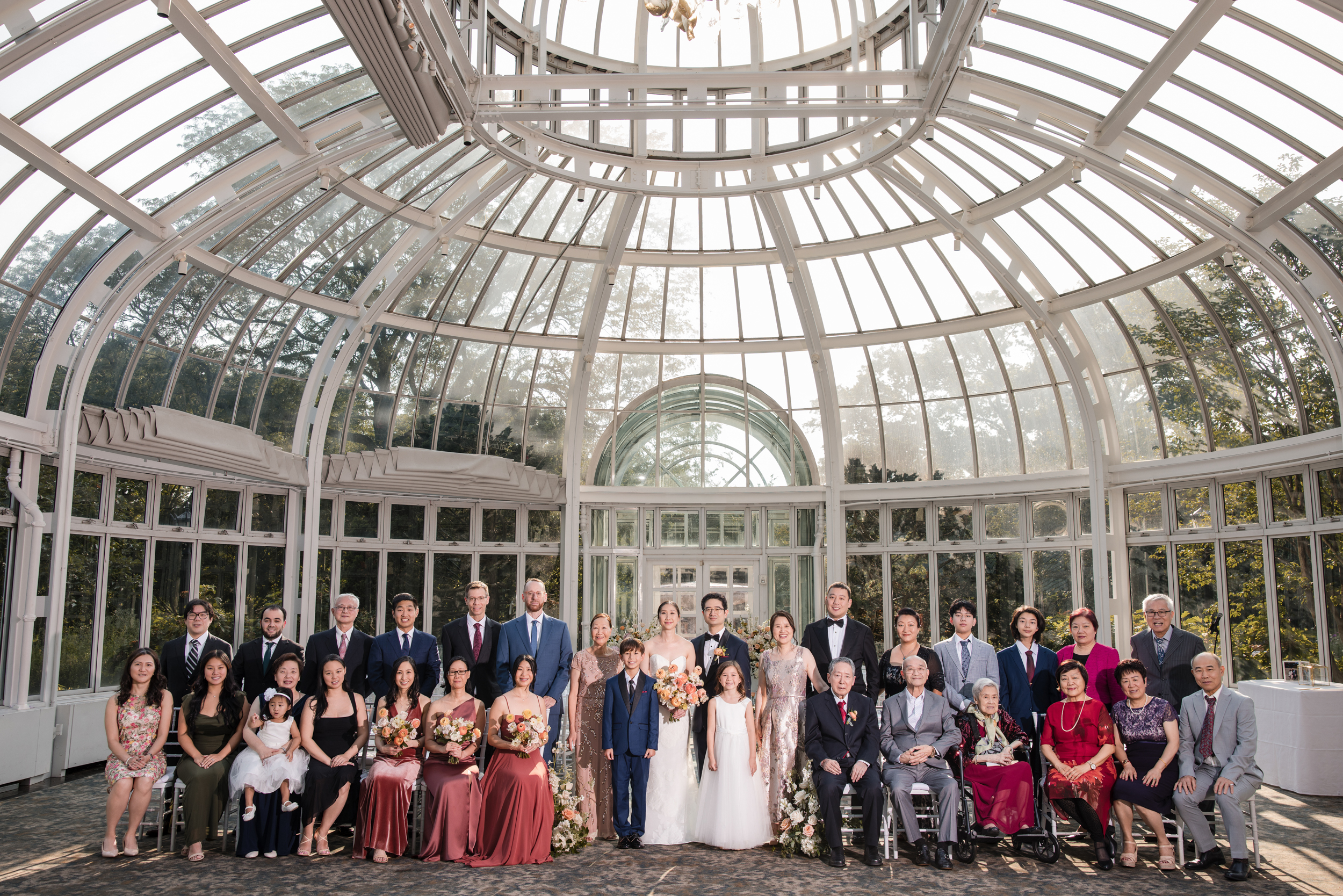 A large group of people, including a bride and groom, pose for a formal photo inside the Brooklyn Botanic Garden Palm House wedding venue, with natural light streaming through the glass conservatory. A large group of people, including a bride and groom, pose for a formal photo inside the Brooklyn Botanic Garden Palm House wedding venue, with natural light streaming through the glass conservatory.