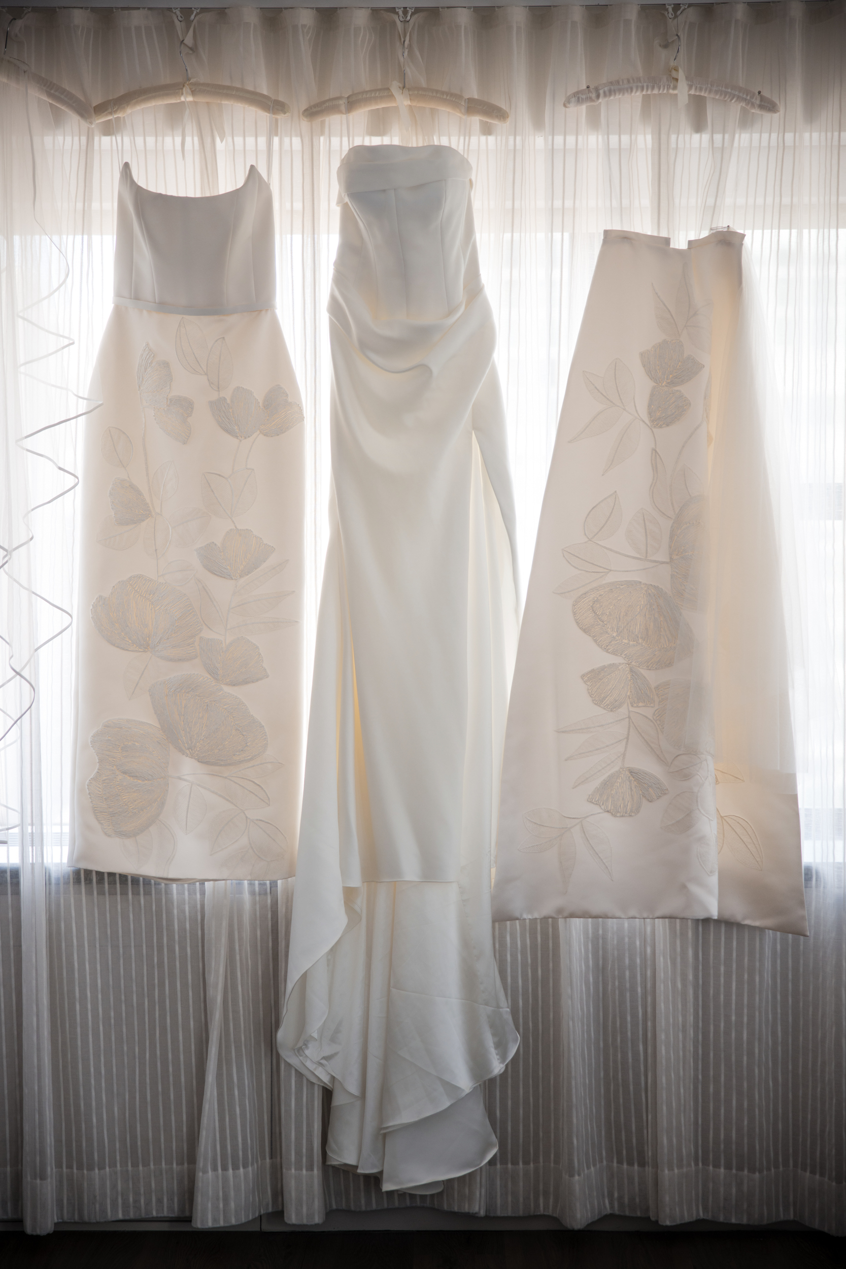 Three white dresses hang in front of a window with sheer curtains; two feature floral embroidery, while the center dress is plain with a structured bodice and draped skirt—perfect for a timeless Lotte New York Palace wedding.