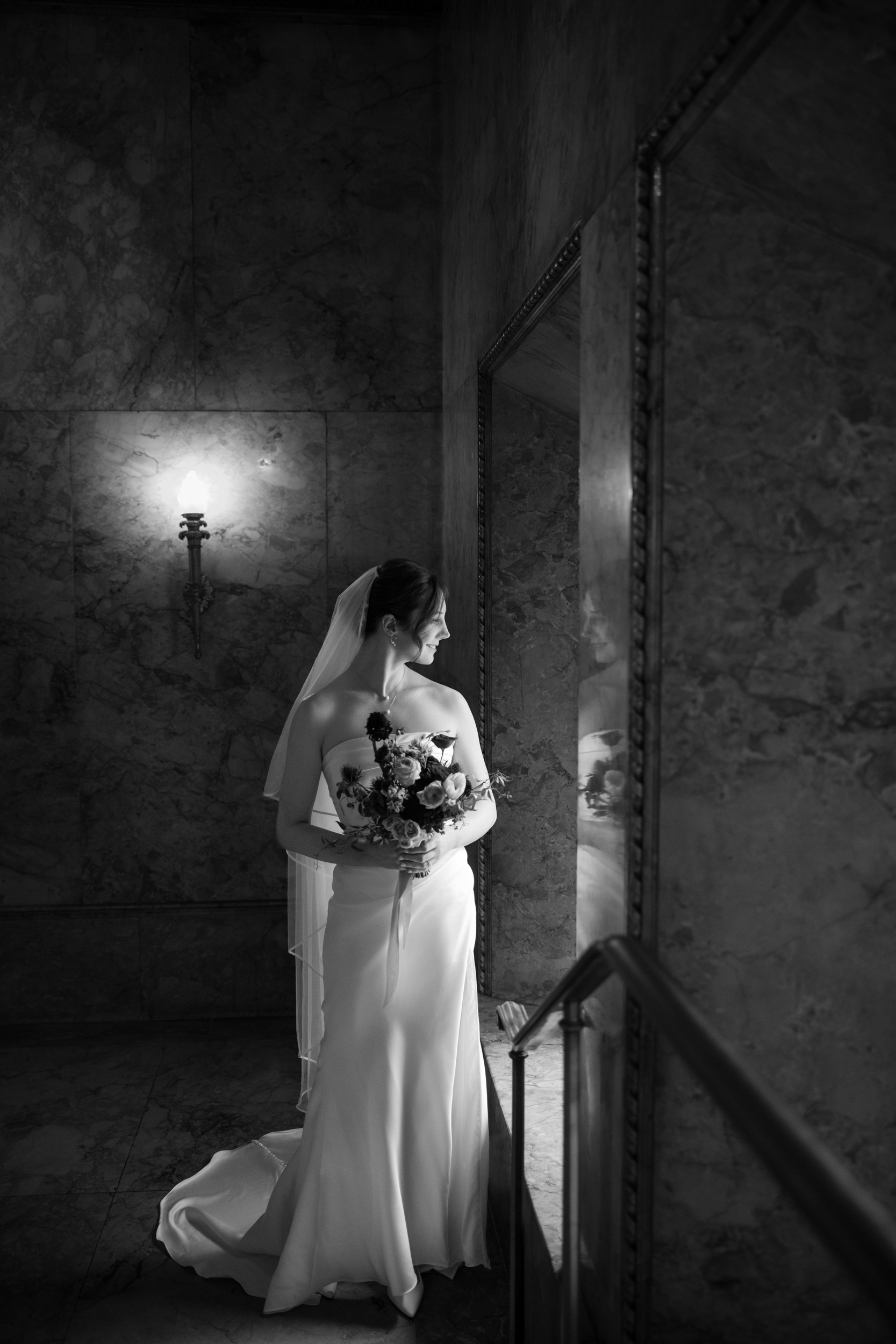 A bride in a strapless gown stands indoors by a window at the Lotte New York Palace wedding venue, holding a bouquet and looking outside. The scene is in black and white, with a marble wall and a lit sconce in the background.