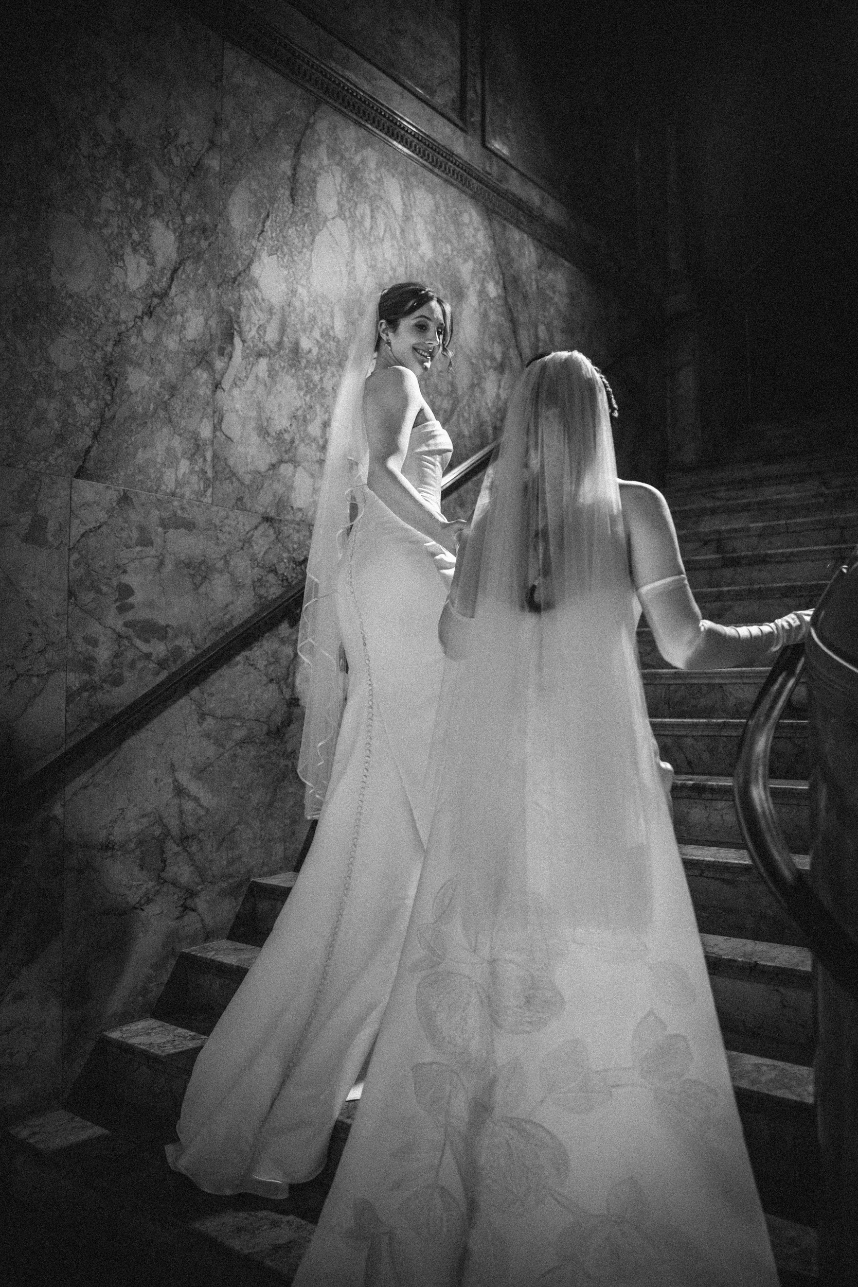 Two women in wedding dresses ascend a marble staircase at the Lotte New York Palace; one looks back over her shoulder while the other faces forward, both wearing long veils.
