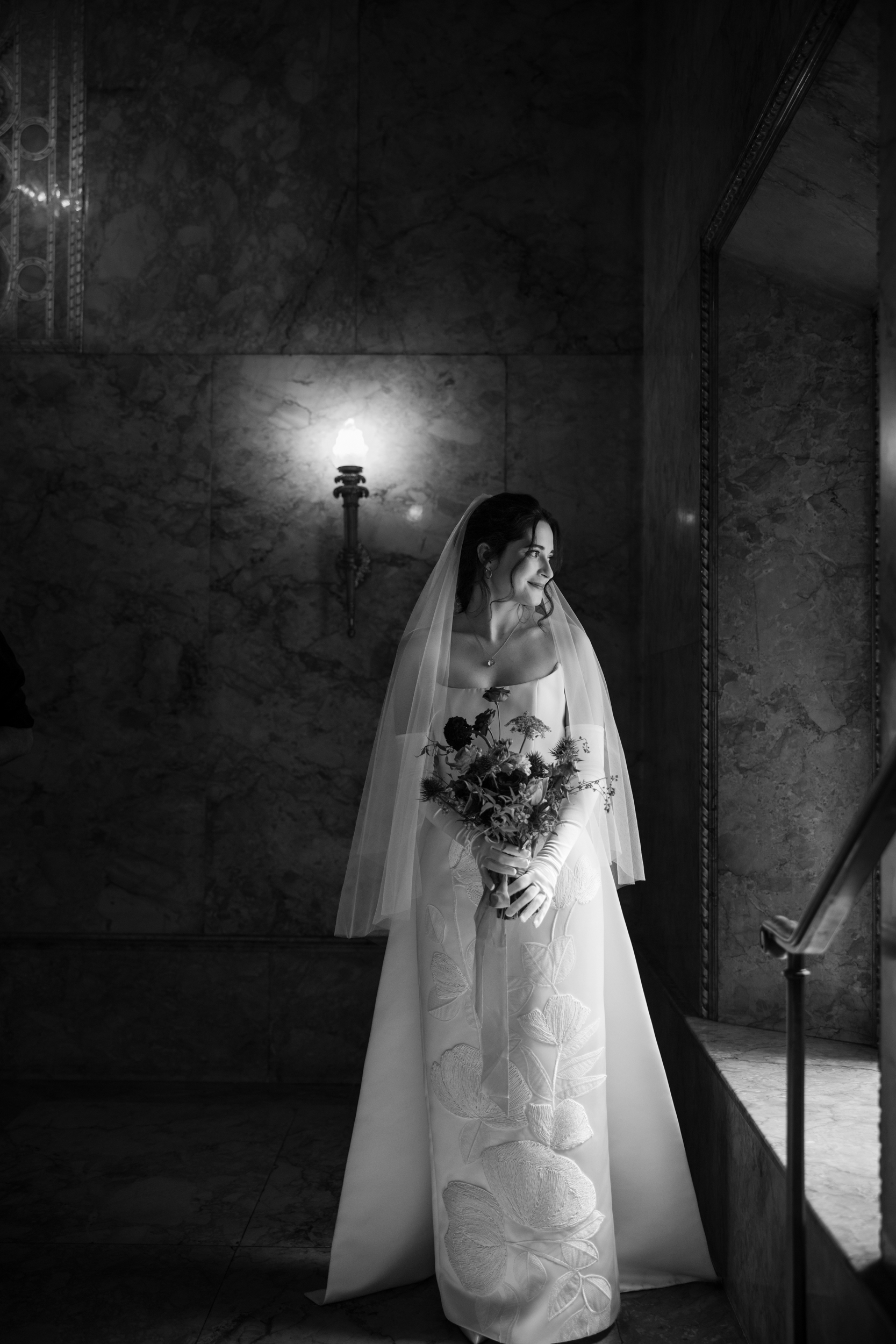 A bride in a white dress and veil stands indoors at her Lotte New York Palace wedding, holding a bouquet and looking out a window, with a lit wall sconce behind her.