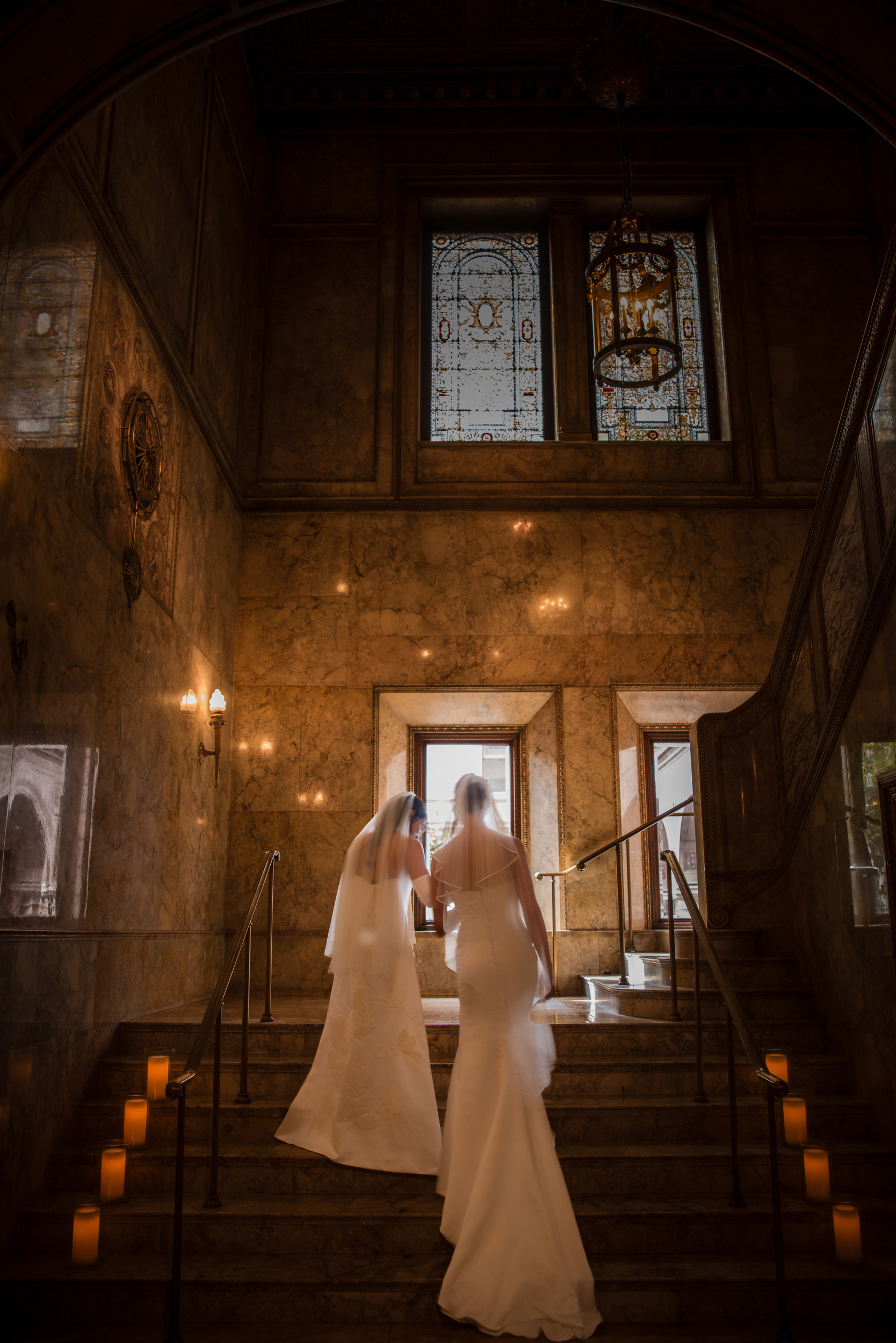 Two women in wedding dresses walk up marble stairs lined with candles inside the elegant, historic Lotte New York Palace, with stained glass windows casting a romantic glow on their wedding day.