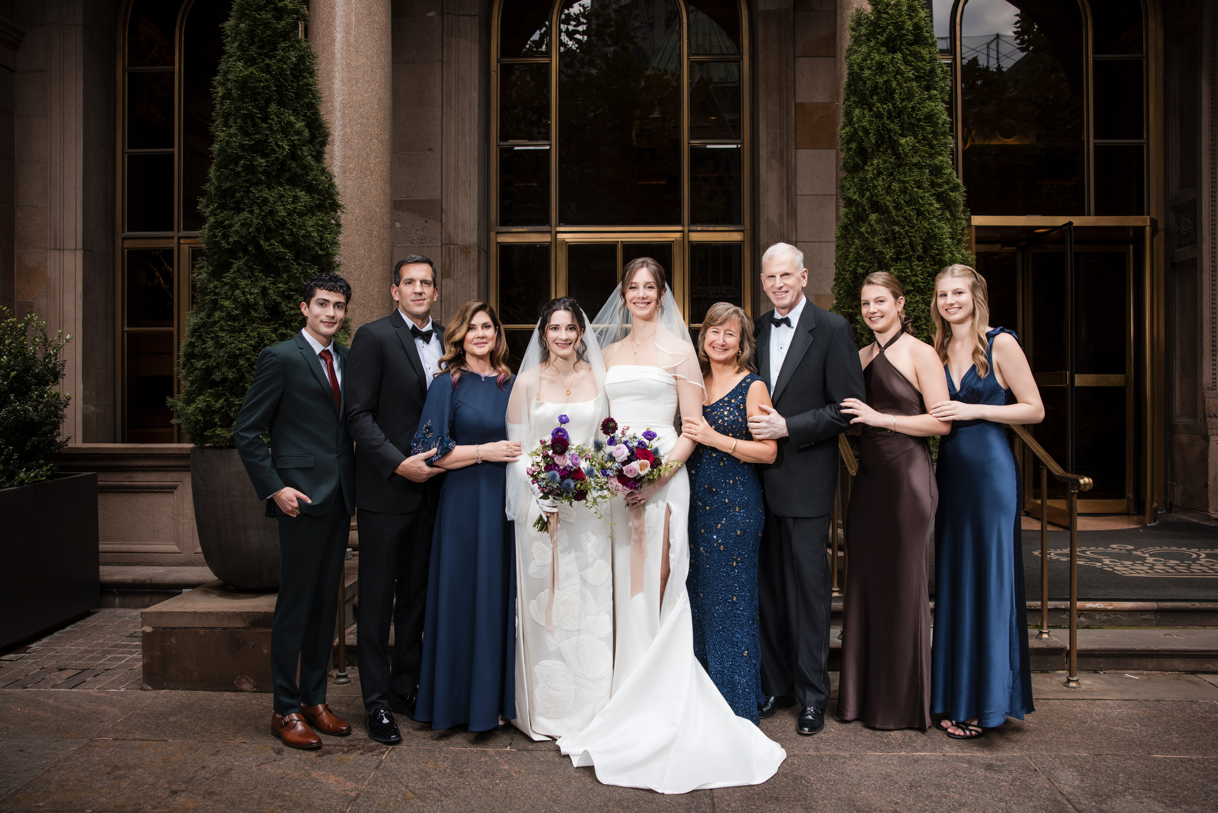 A wedding party poses outside the Lotte New York Palace; two brides stand in the center in white, surrounded by family and friends in formal attire.