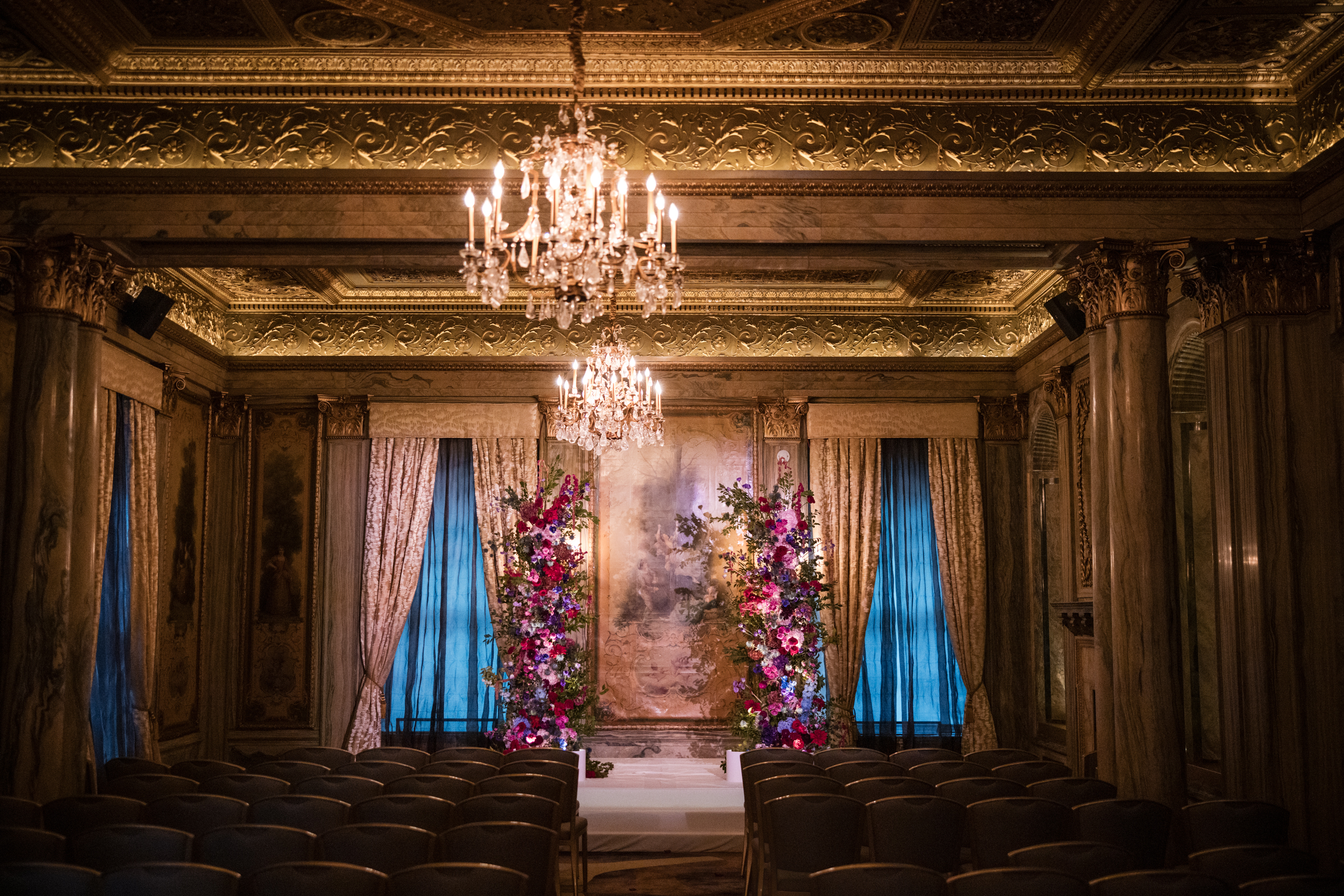 Ornate gold room with chandeliers, rows of chairs, and a flower-adorned altar at the front—this elegant setting, framed by tall blue curtains and marble columns, captures the timeless romance of a Lotte New York Palace wedding.