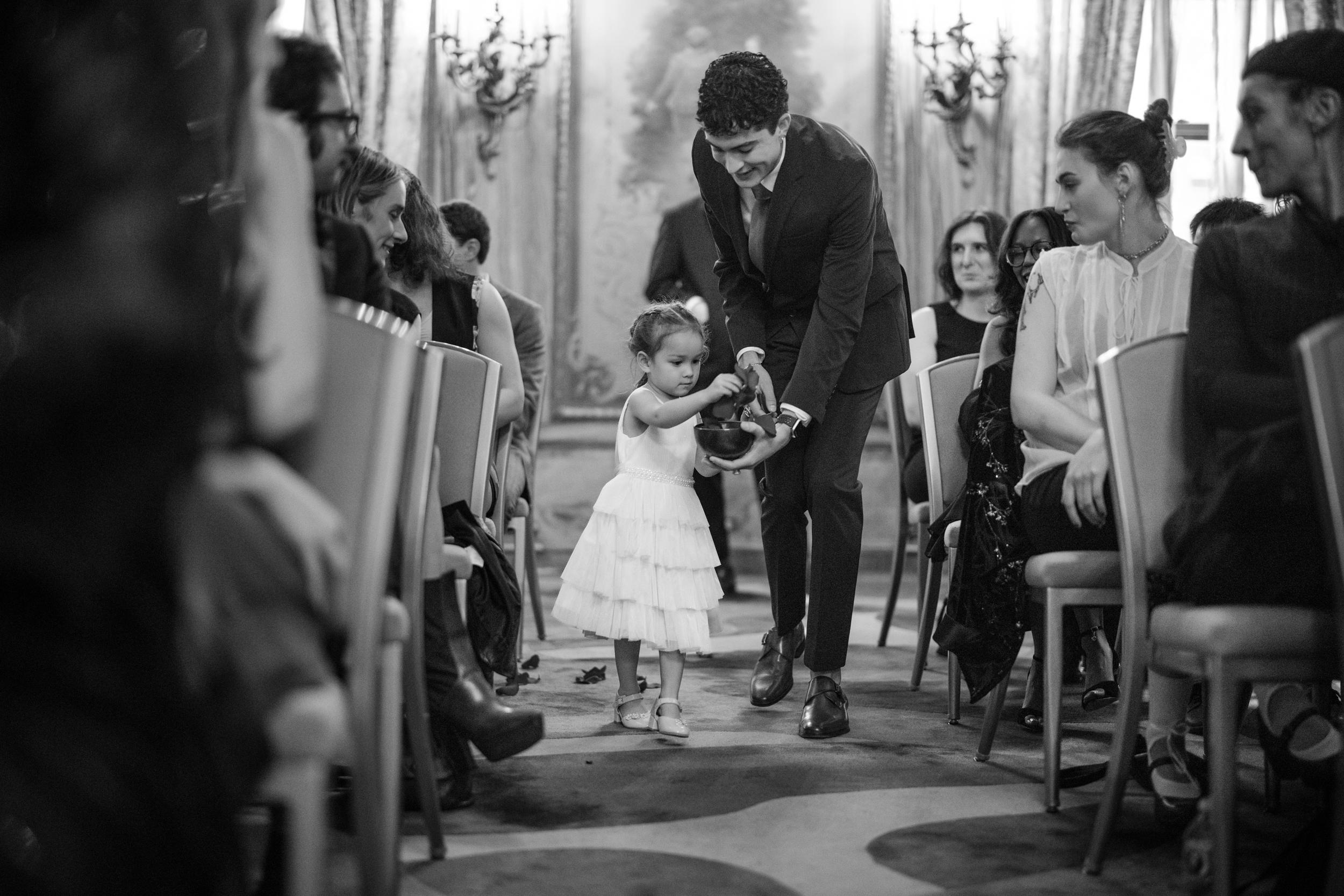 A man helps a young girl in a white dress walk down the aisle during a formal event at a Lotte New York Palace wedding as seated guests look on.