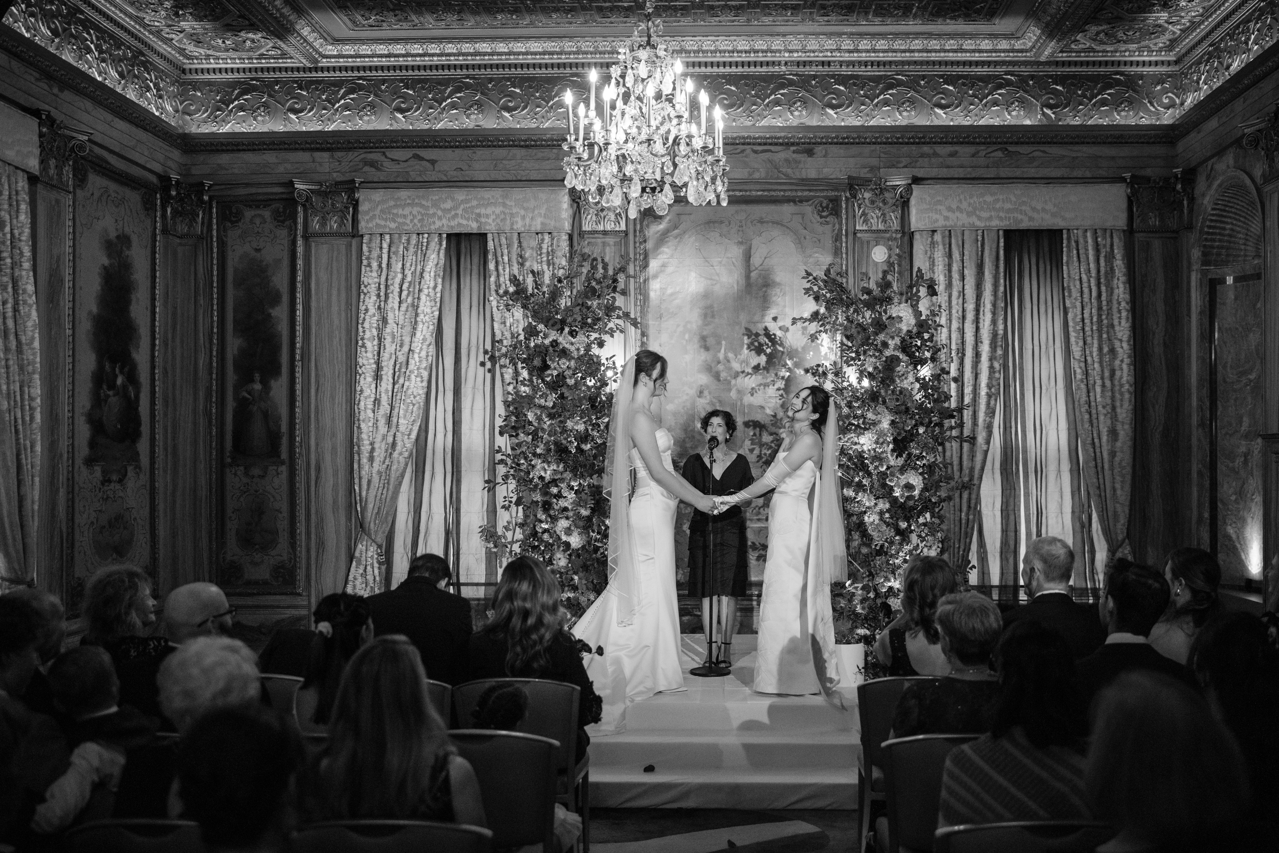 Two brides in white dresses stand facing each other, holding hands during a wedding ceremony in an ornate, chandelier-lit room at the Lotte New York Palace, with guests seated and watching.
