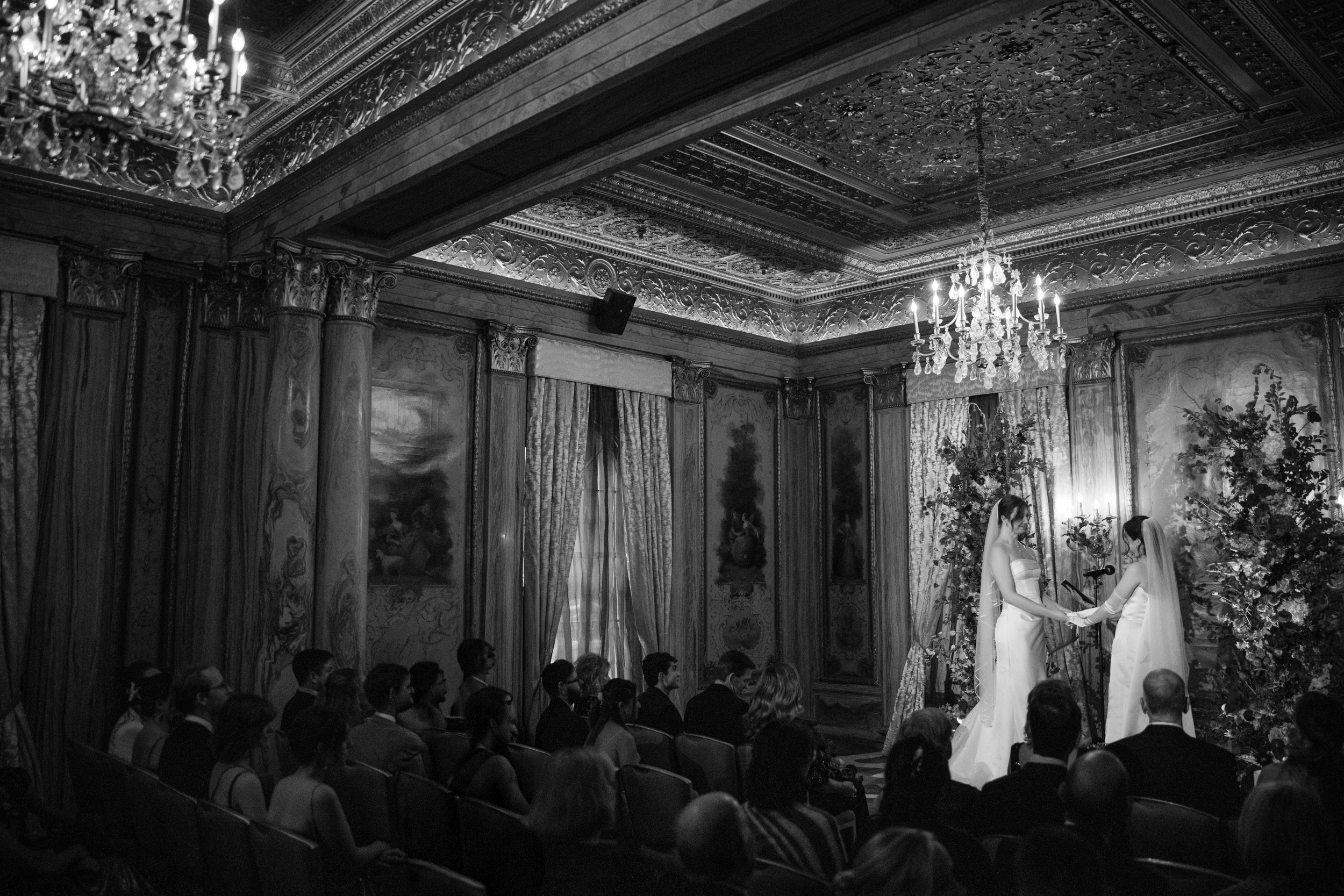 Two brides in wedding attire stand at the altar holding hands in an ornate, chandelier-lit room at a gorgeous Lotte New York Palace wedding as guests watch the ceremony.