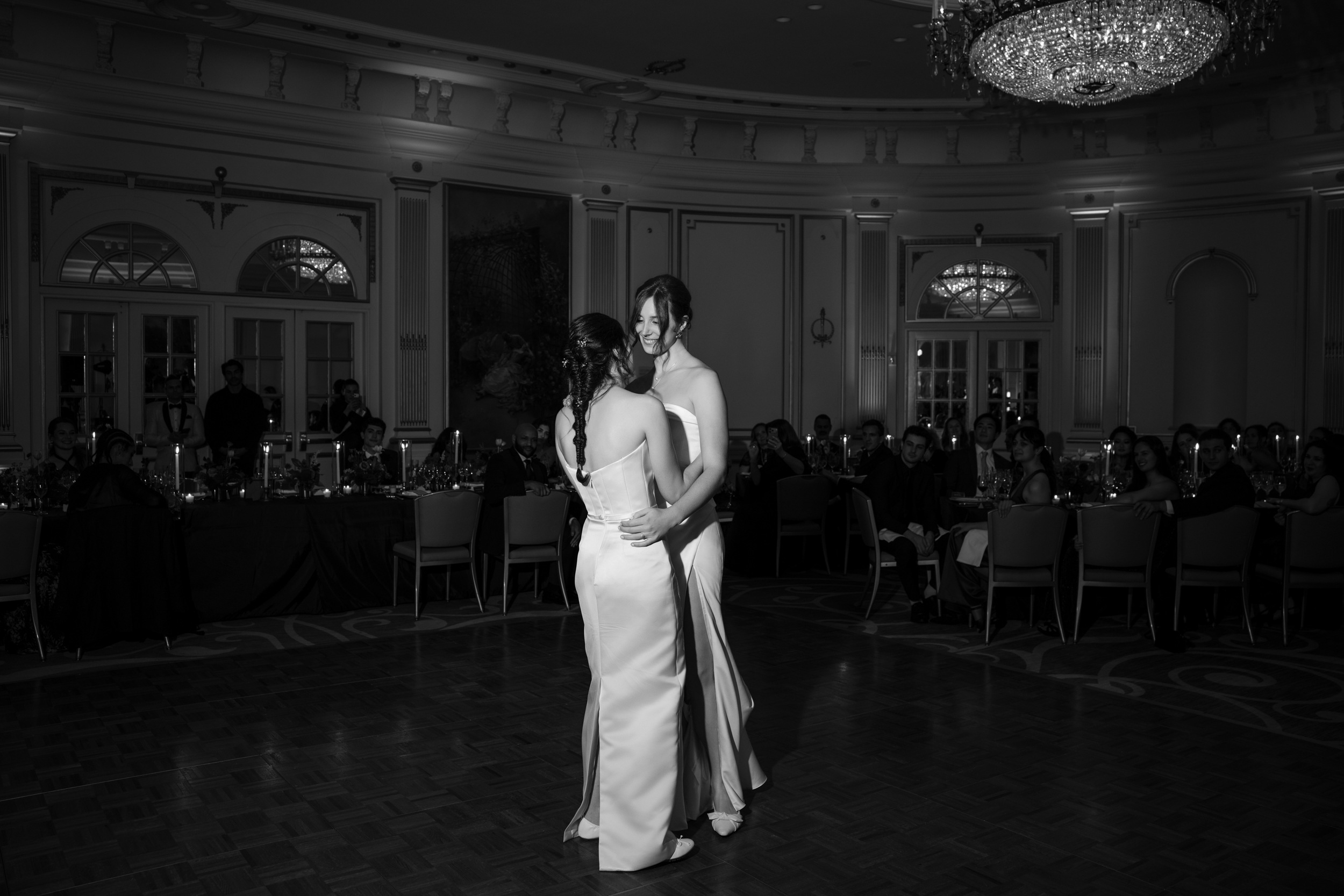 Two women in formal gowns dance together in the center of an elegant, chandelier-lit ballroom at a Lotte New York Palace wedding, as guests watch from seated tables.