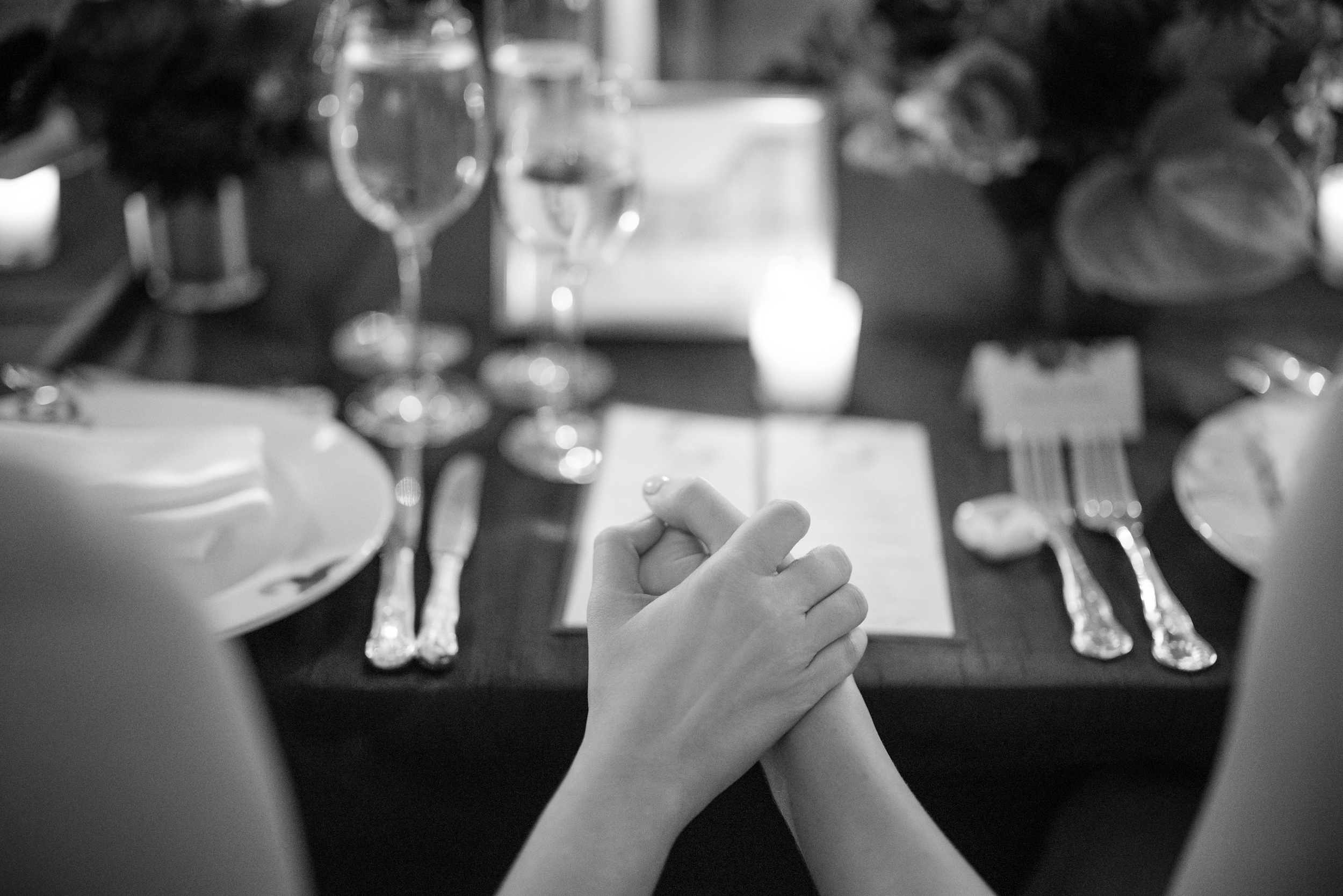 Two people hold hands across a formally set dinner table with plates, silverware, wine glasses, and a lit candle—capturing an intimate moment reminiscent of a Lotte New York Palace wedding.