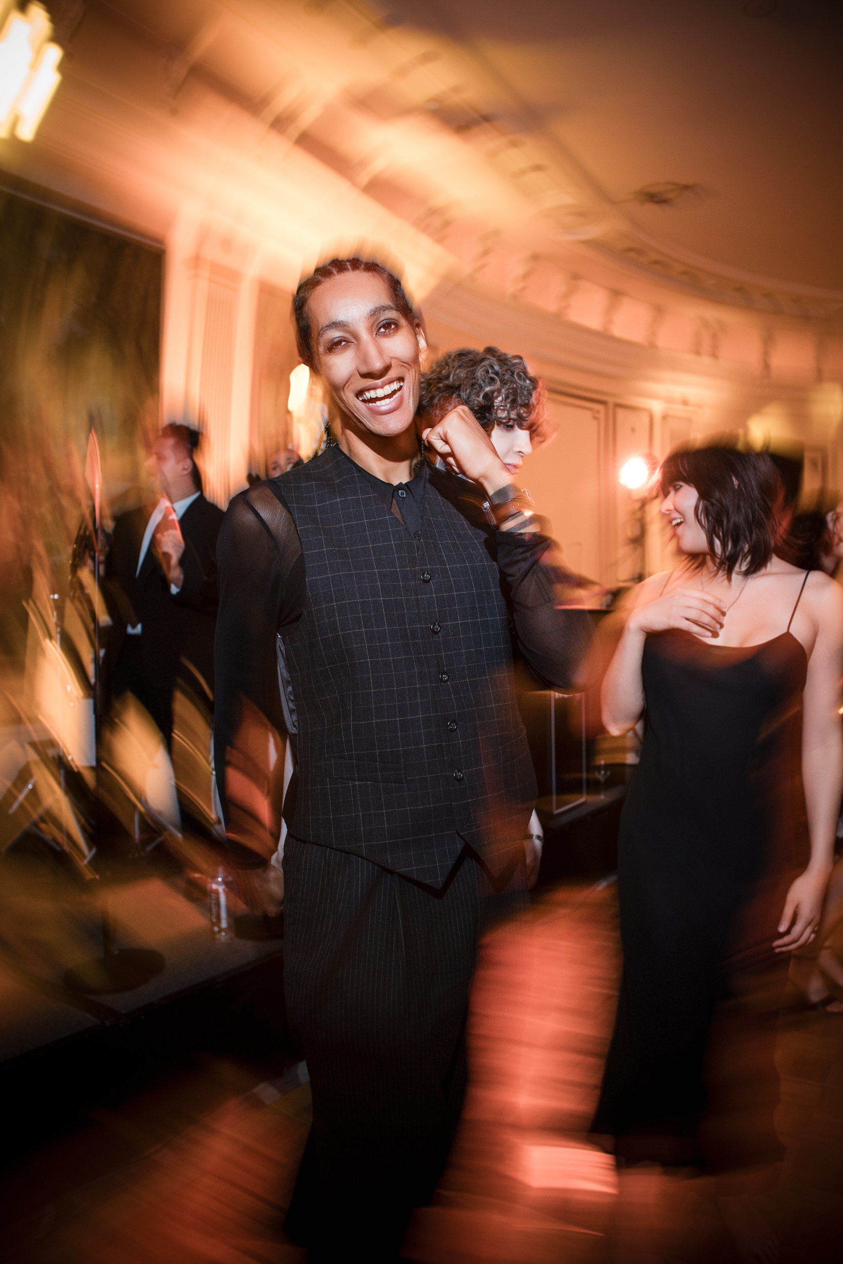 A person in a dark suit smiles at the camera on a dance floor at a Lotte New York Palace wedding, with blurred partygoers and warm lighting in the background.