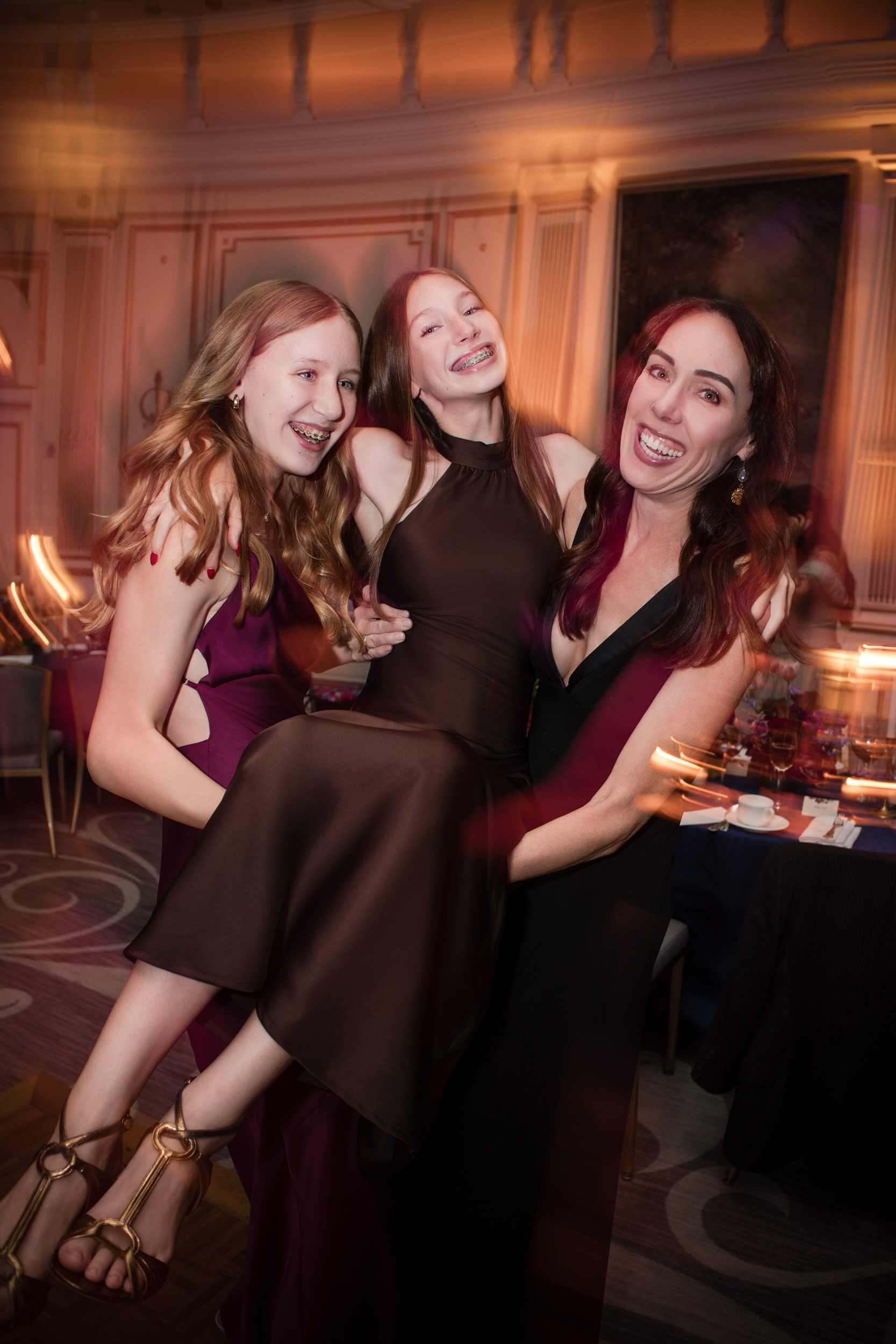 Three people in formal attire smile and pose together at an indoor event, with one young girl being lifted by the others. The background shows a decorated, elegant room reminiscent of a Lotte New York Palace wedding.