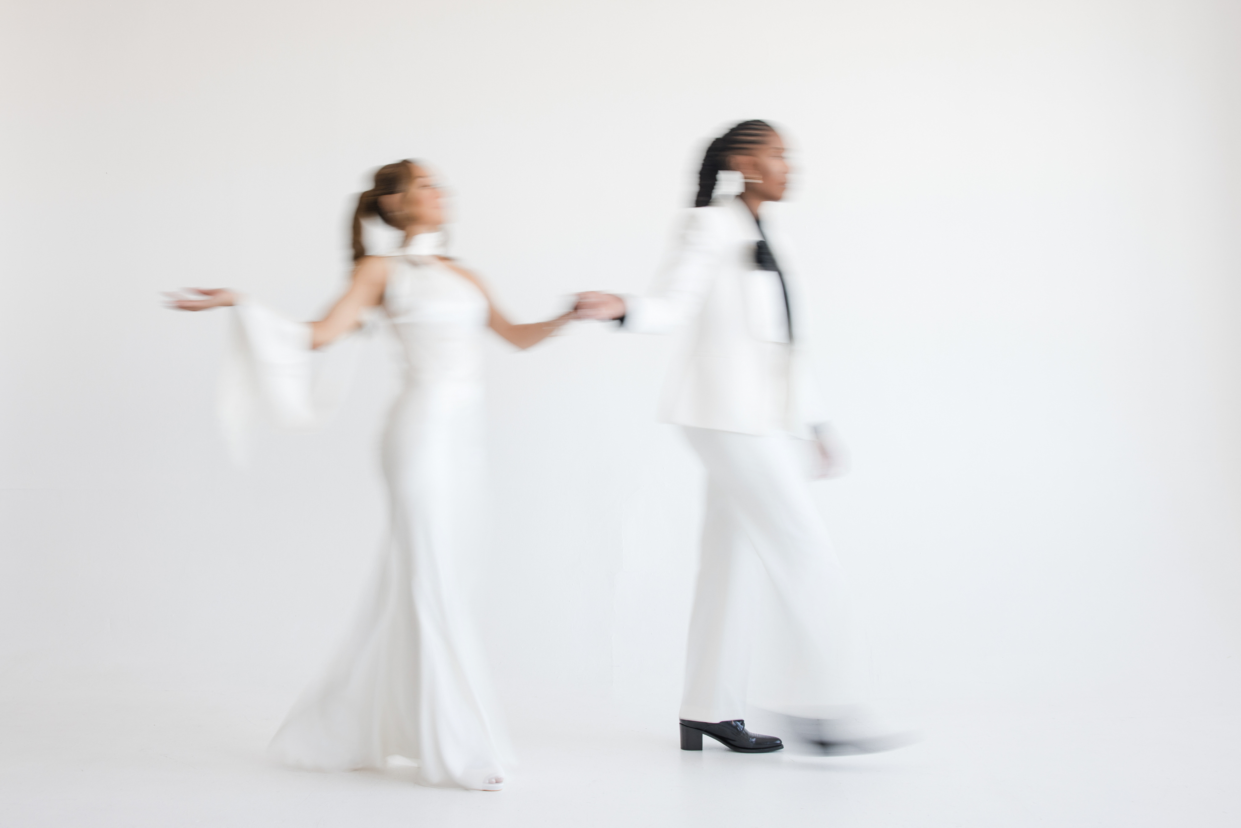 Two people in white formal outfits hold hands while walking; the image is intentionally blurred, giving a sense of motion against a plain white background, capturing the modern romance of an NYC studio engagement session.