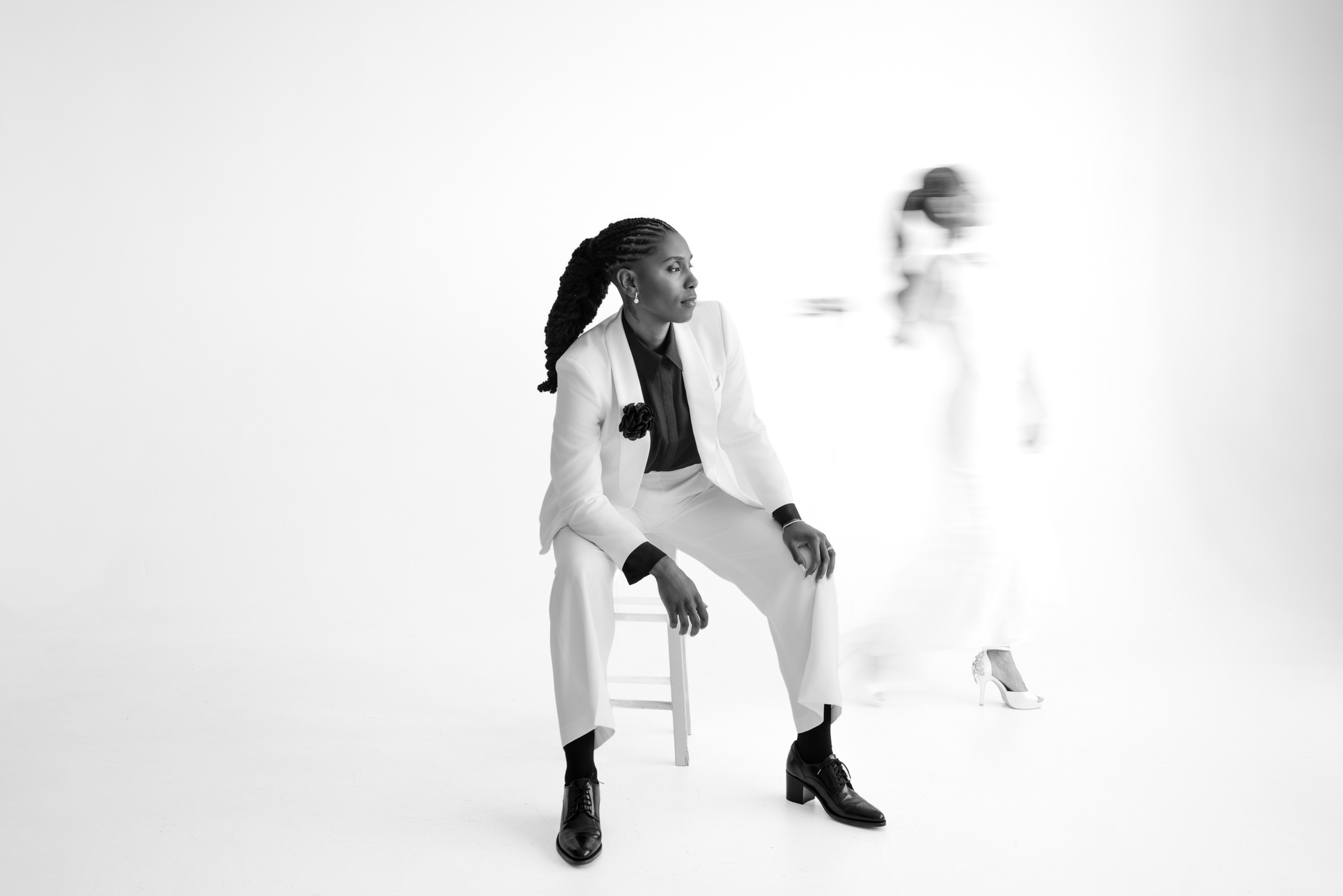 Person in a white suit sits on a stool while another person in a white dress walks by, blurred, against a white background—capturing the modern elegance of an NYC studio engagement session.